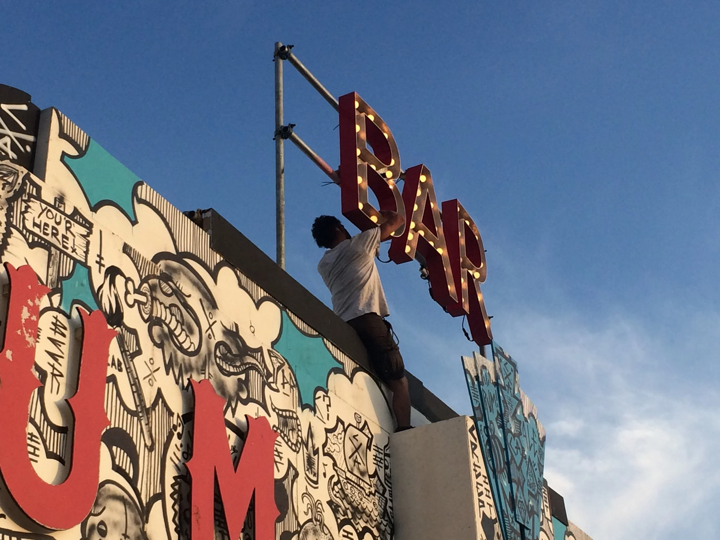 Menschen installieren ein leuchtendes Signet an einem Gebäudeteil bei Dämmerung, mit Fantasiewolken-Graffiti im Hintergrund.