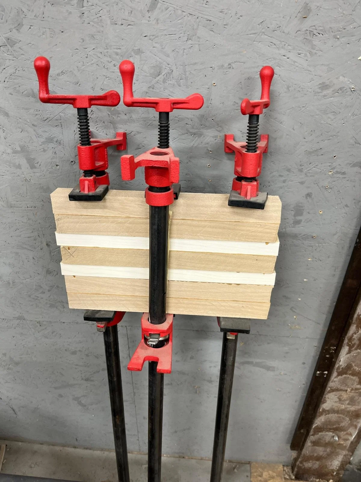 Drei rote Schraubzwingen, die ein Stück Holz auf einer Werkbank halten.