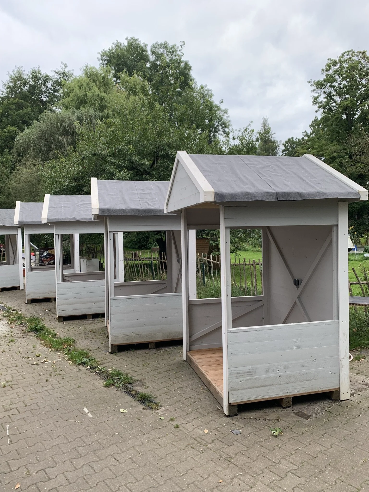 Mehrere kleine weiße Hundehütten mit grauen Dächern stehen auf gepflastertem Boden in einem Park, im Hintergrund sind Bäume und ein bewölkter Himmel.