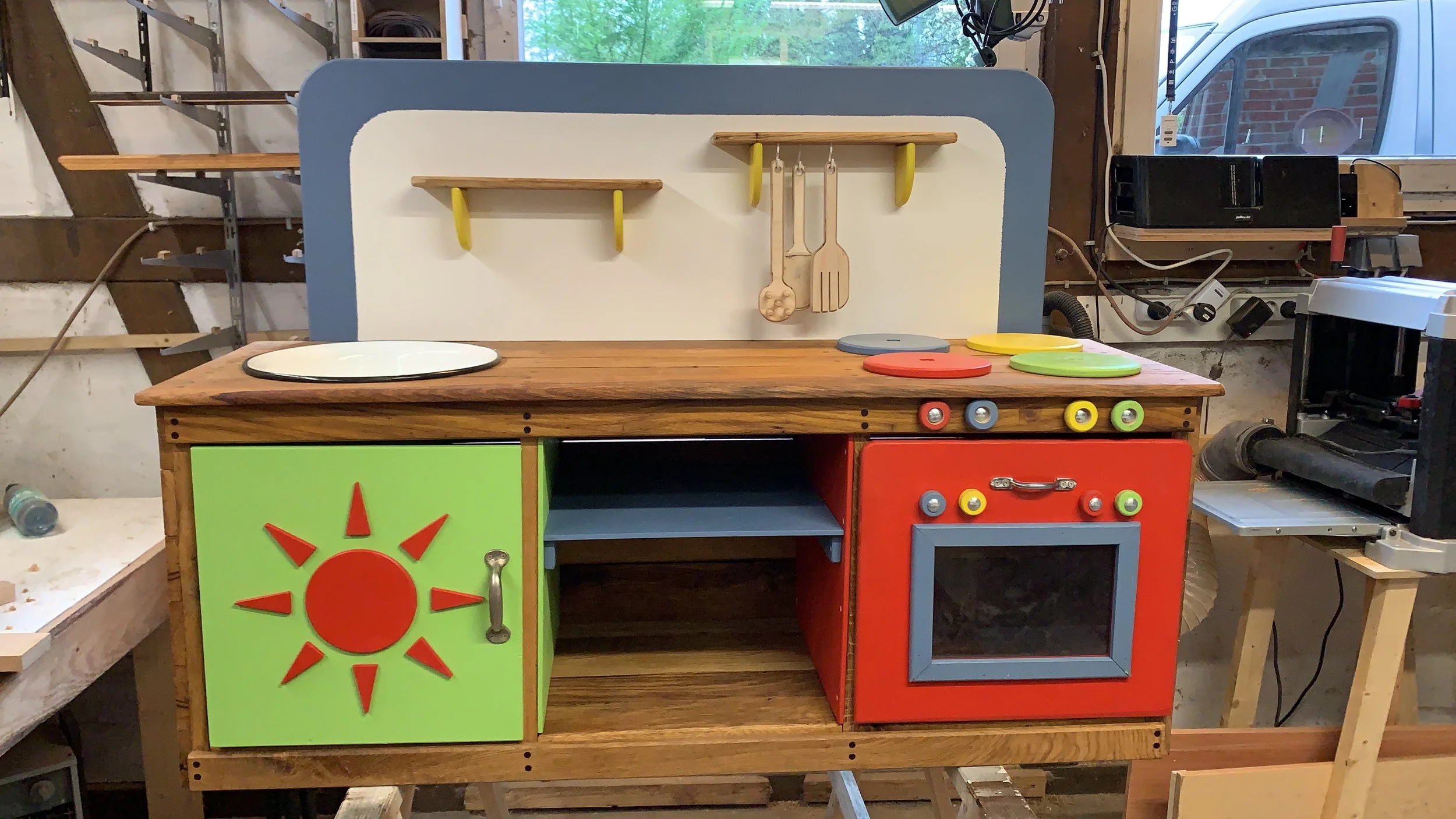 Holzküche für Kinder mit bunten Türen, einer weißen Herdplatte, Holzgabel und Löffel, auf einem Holzposten im Spielzimmer.