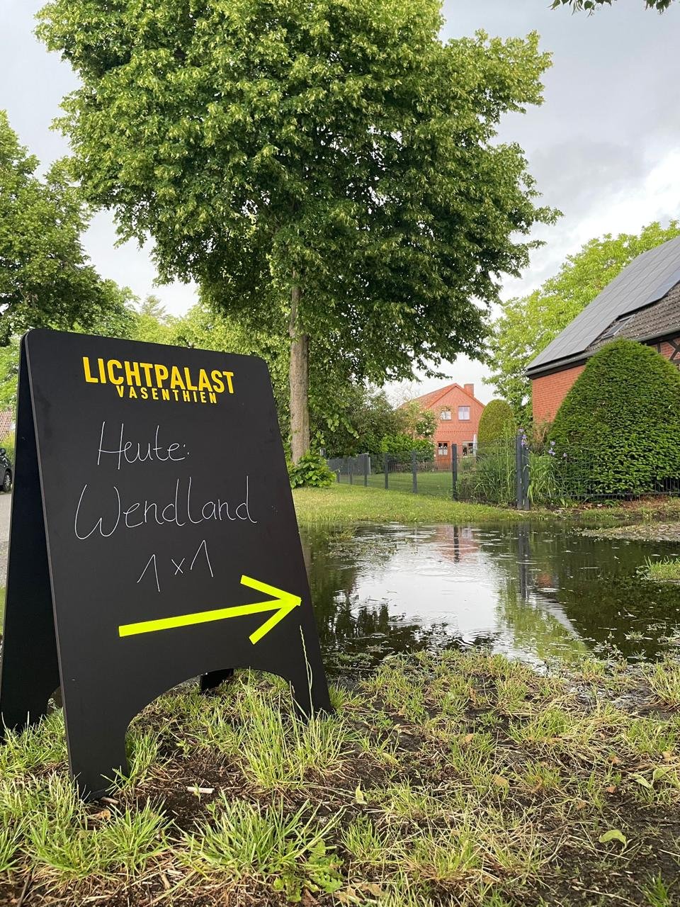 Ein Straßenwerbeschild mit schwarzer Oberfläche und gelber Schrift, das auf einen nahegelegenen Wasserstau oder Pfütze hinweist, umgeben von grünem Gras und Bäumen, mit Häusern im Hintergrund bei bewölktem Himmel.
