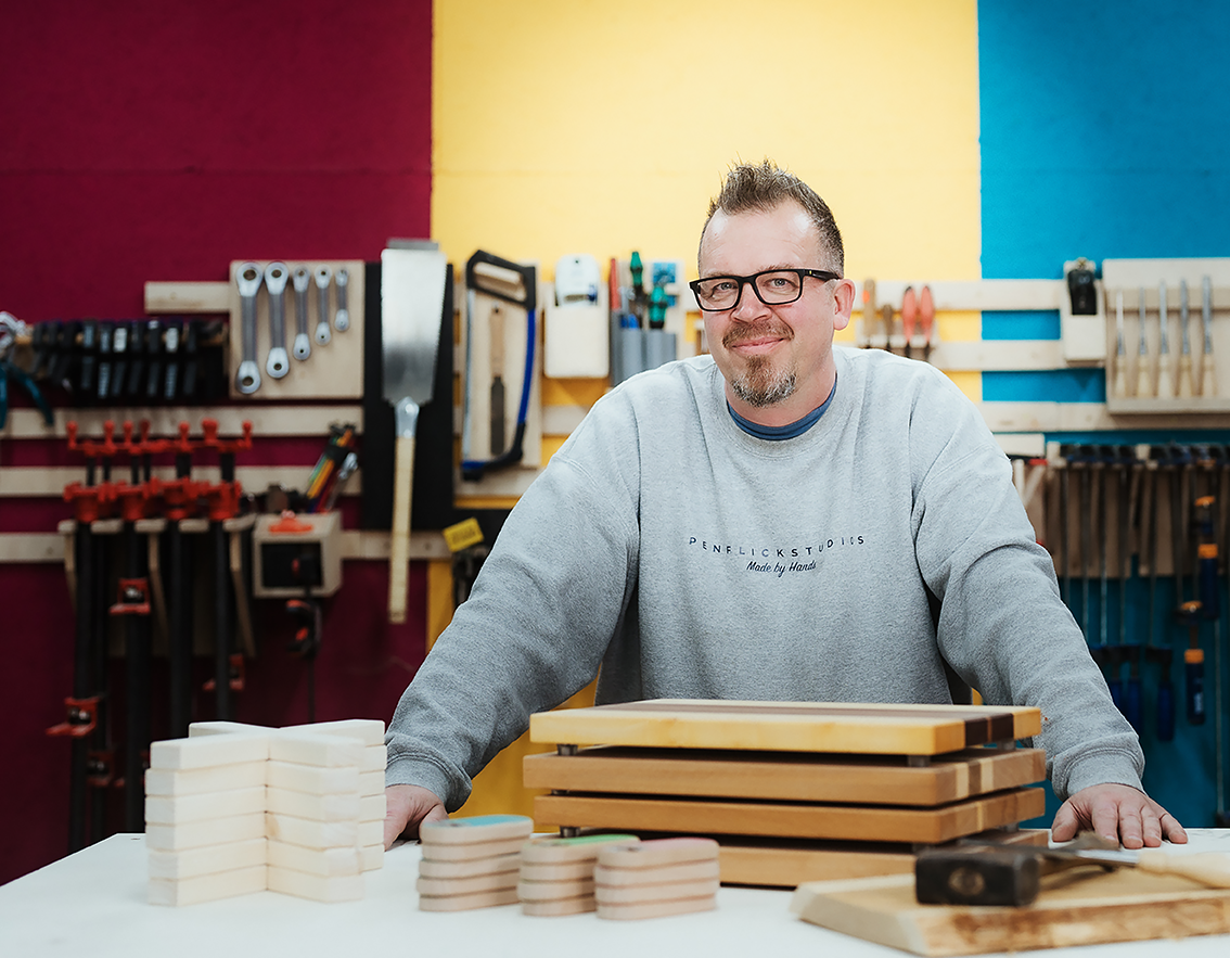 Ein Mann mit Brille und grauem Sweatshirt steht in einer Werkstatt vor einem Tisch mit Holzstücken, Werkzeug und kleinen Holzscheiben. Die Werkstatt ist bunt gestrichen mit roten, gelben und blauen Flächen im Hintergrund.