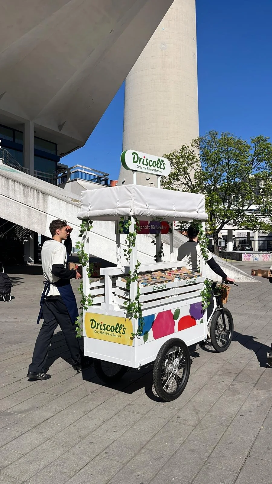 Mobiler Obststand mit der Marke Driscoll's, verkauft Beeren, im Freien bei sonnigem Wetter, mit zwei Personen in der Nähe, eine Person schiebt den Stand, die andere fährt Fahrrad.