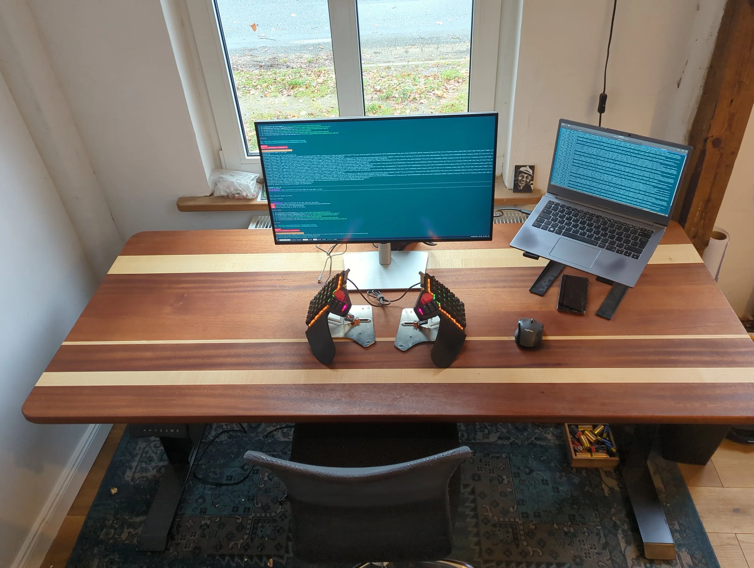 Ein Arbeitsbereich mit einem großen Monitor, einem Laptop auf einem Ständer, einem drahtlosen Lautsprecher, einem Smartphone, zwei Tastaturen, einer Maus, einem Glas mit Batterien, einem kleinen Bild und einem Kabel, vor einem Fenster mit Blick auf d