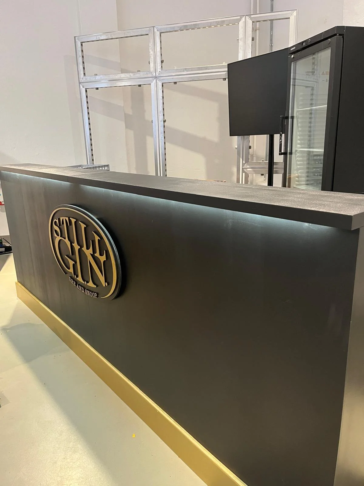 Ein Schwarz-grauer Tresen mit einem Logo, auf dem steht »SILVER GUN« und darunter »DRE AND SNOOP«. Im Hintergrund ist ein metallener Rahmen und ein Kühlschrank mit Glasfront zu sehen.