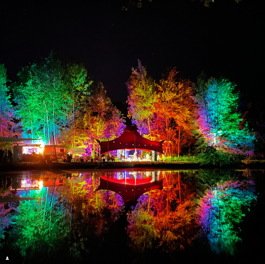 Lichter erleuchtete Bäume und eine Zeltstruktur an einem See bei Nacht, mit deren Spiegelung im Wasser.