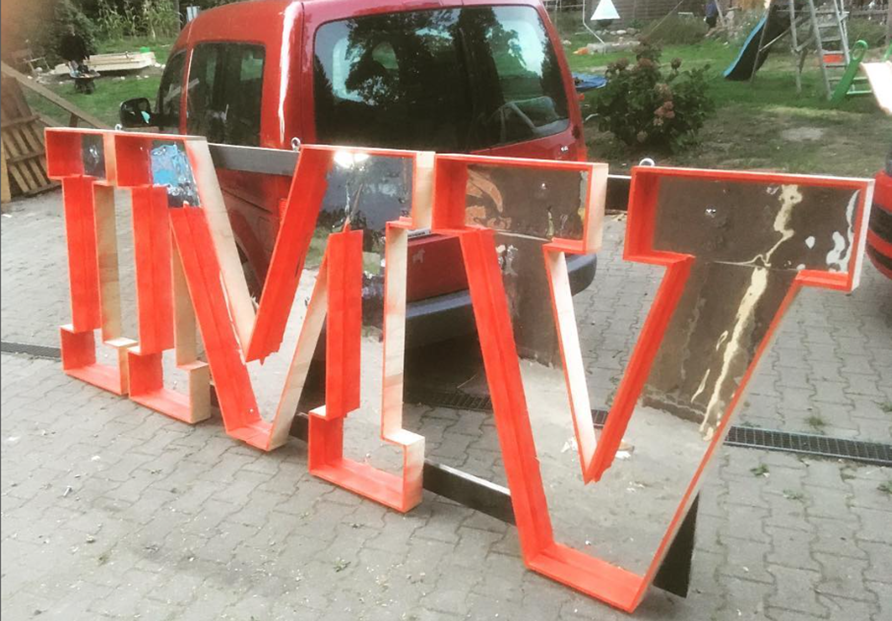Großer Schriftzug mit leeren Rahmen, wahrscheinlich für eine Schau- oder Werbetafel, vor einem roten Auto auf gepflastertem Boden in einem Garten mit Spielgeräten und Pflanzen.