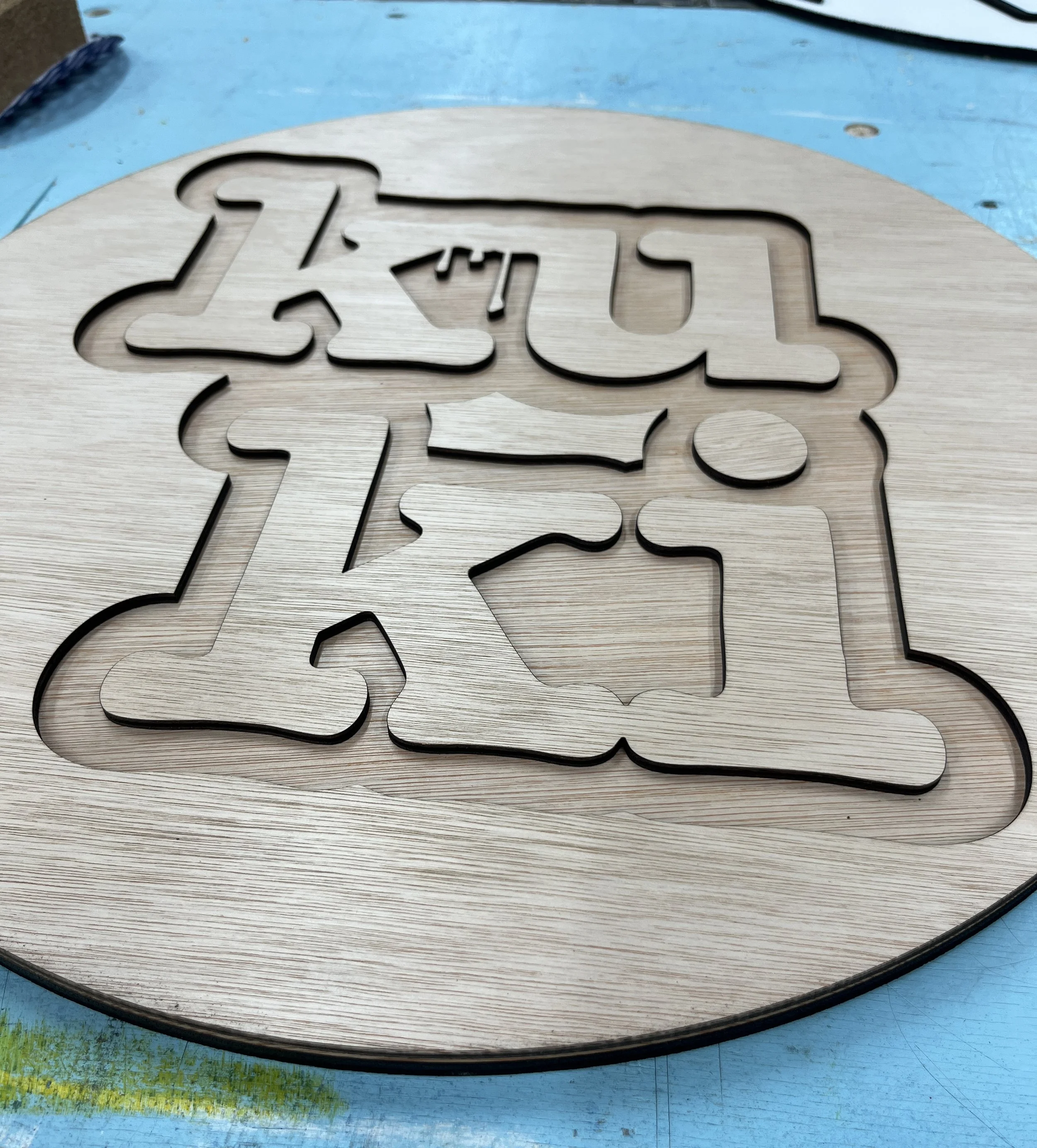 Ein Holzschild mit horizontal eingeschnittenen Buchstaben. Die Buchstaben sind teilweise sichtbar, es erscheinen die Buchstaben 'z', 'u', 'g', 'e', 's'. Das Schild liegt auf einer blauen Holzplatte, die an einer Ecke grünlich ist.