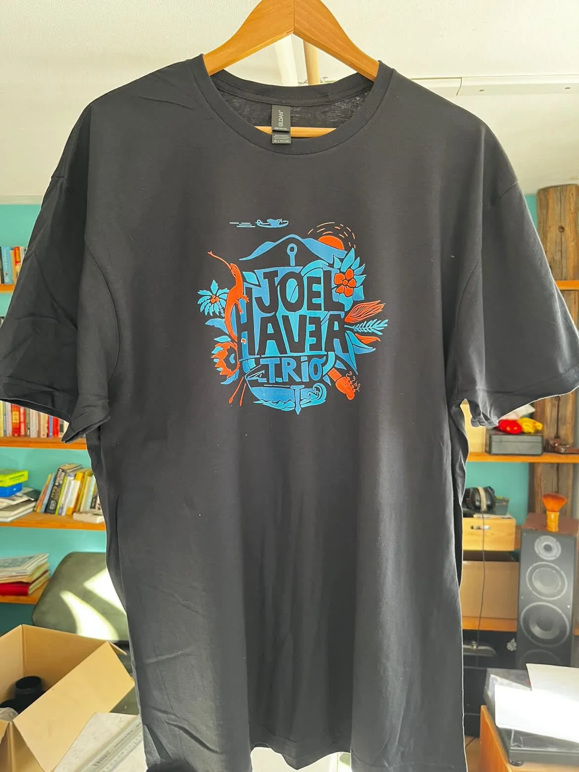 Schwarzes T-Shirt mit grafischem Design und Text, auf einem Holzkleiderständer. Hintergrund mit Büchern, einem Regal und anderen Gegenständen.