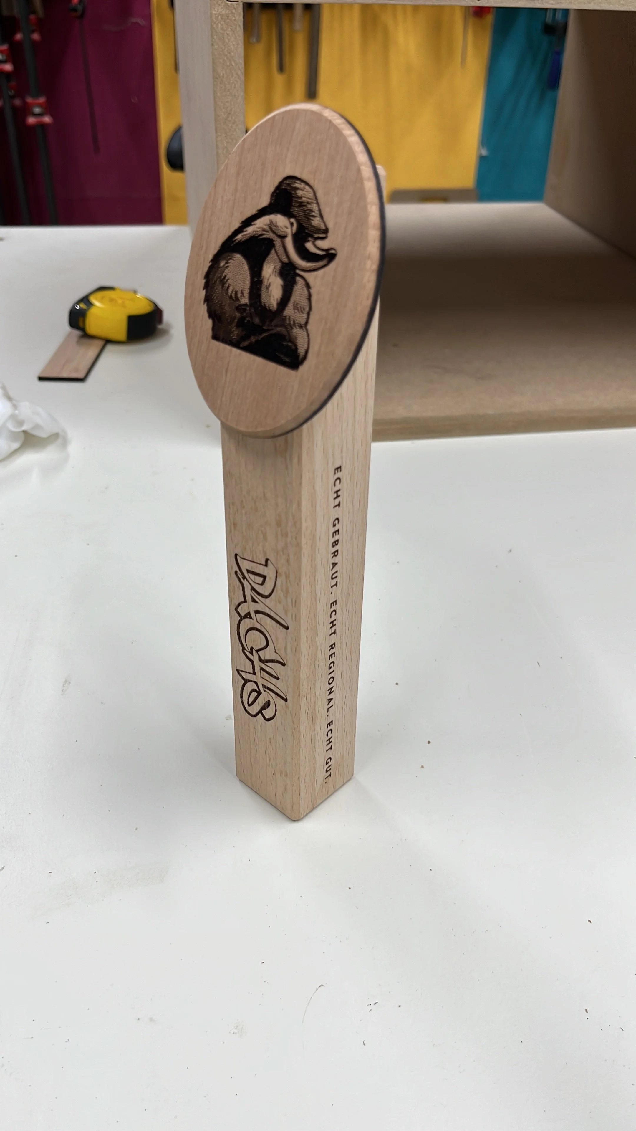 Holzspielzeug mit Elefantenmotiv und Text in deutscher Sprache auf der Seite, auf weißem Tisch, Hintergrund mit bunten Werkzeugen.