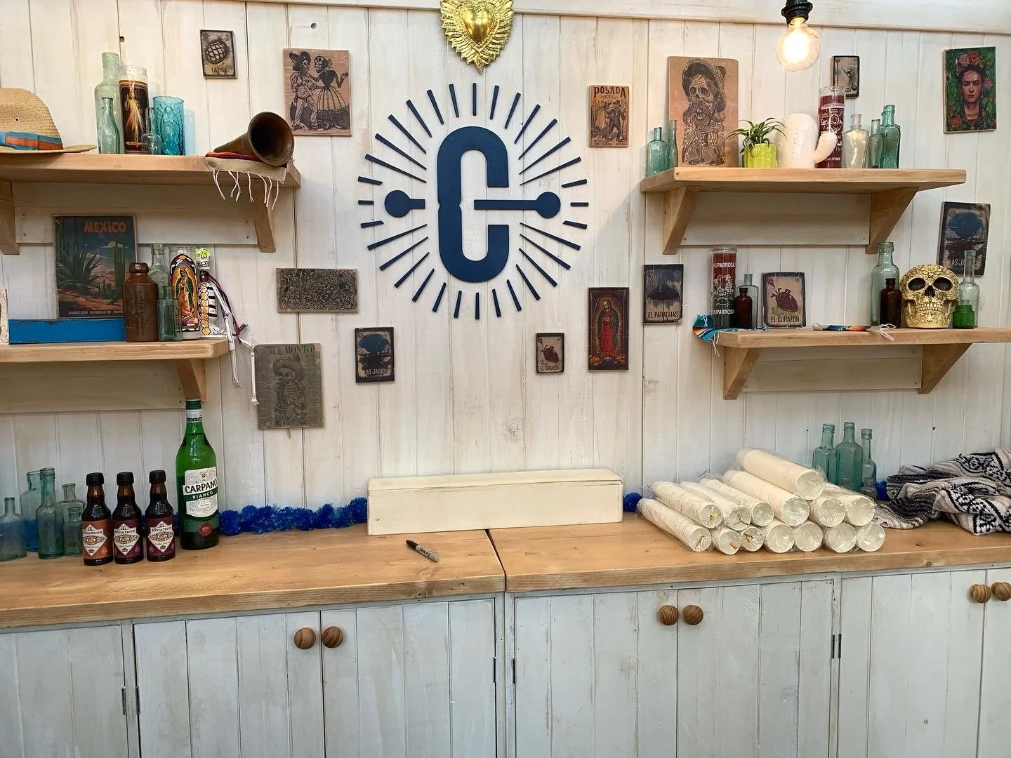 Inneneinrichtung mit Wanddekorationen, darunter Wandkunst mit einem großen G-Logo, Regale mit Flaschen, Skulpturen und Dekorationen, sowie Holzschränke und eine Arbeitsfläche mit Getränkeflaschen und Papierrollen.