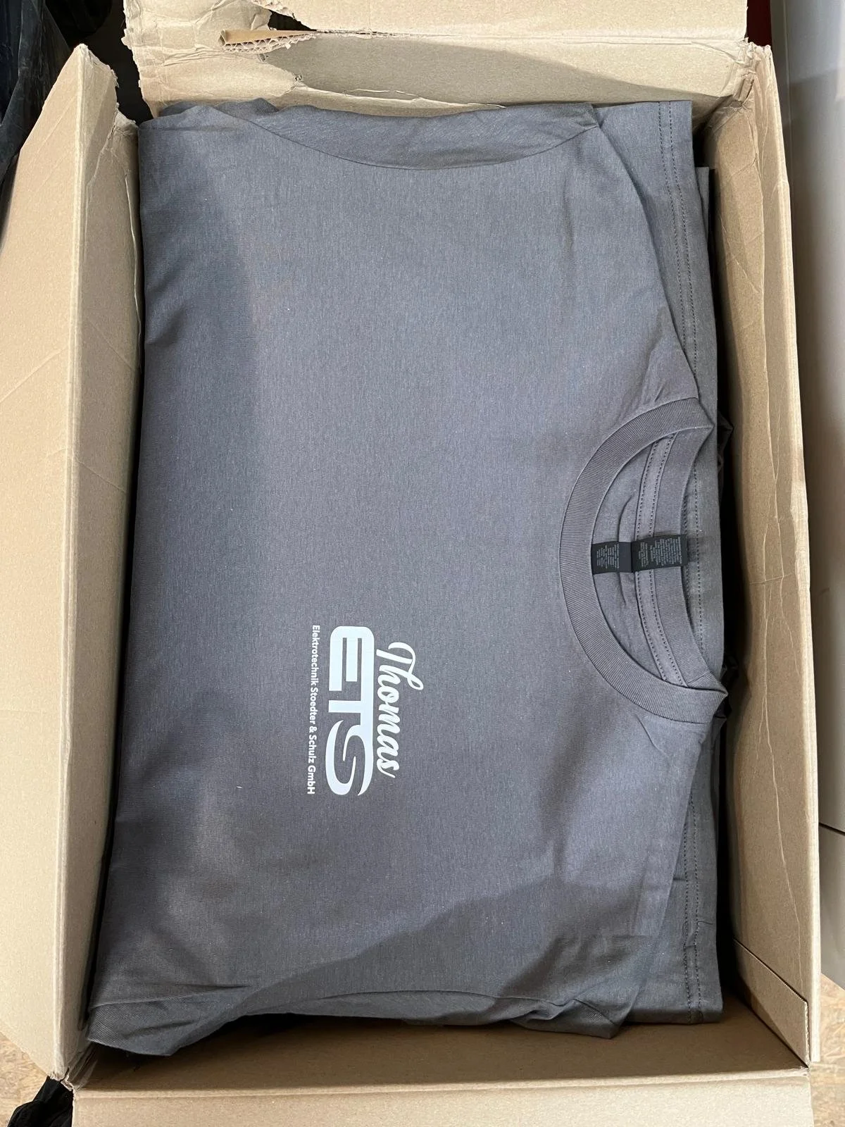 Graues T-Shirt mit weißem Logo, verpackt in einer Pappschachtel.