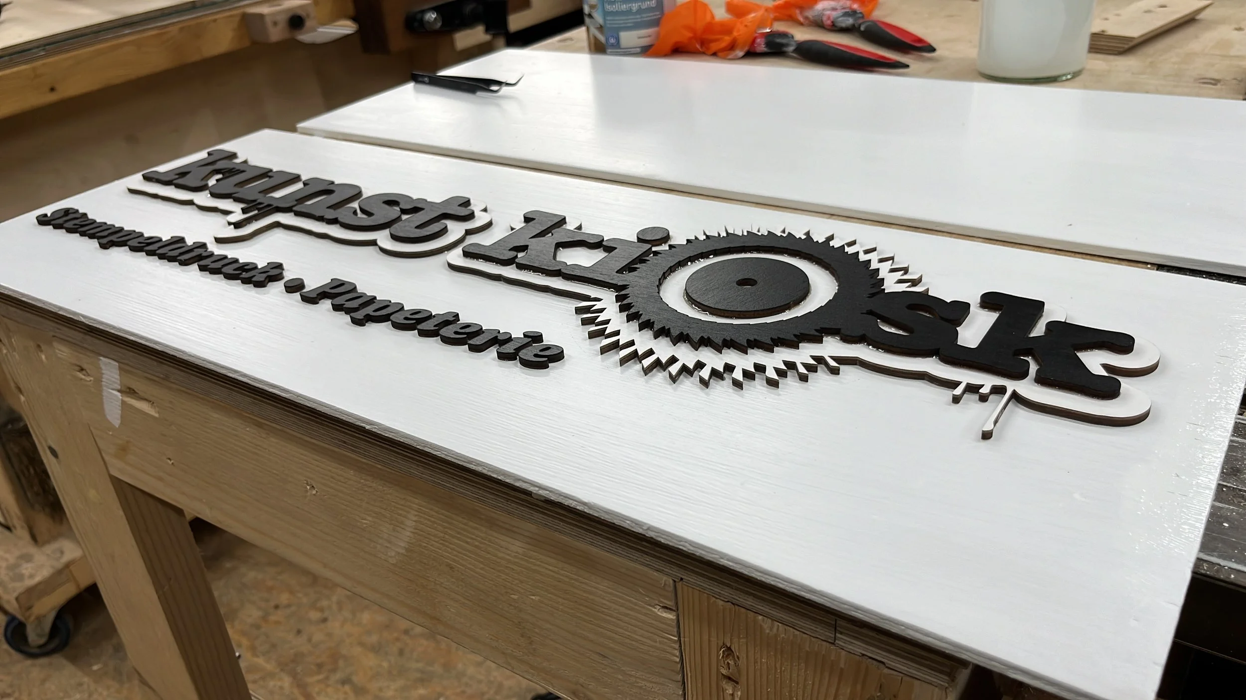 Ein Holzschild mit schwarzer gravierter Schrift und einem Zahnrad-Design. Das Schild liegt auf einem weißen Untergrund auf einem Holztisch. Im Hintergrund sind Werkzeuge und Materialien sichtbar.