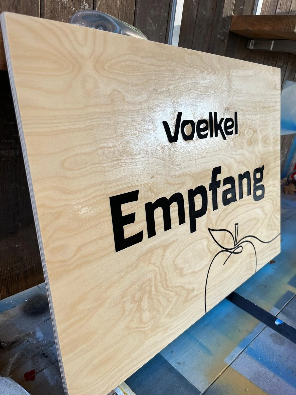 Ein Holzschild mit schwarzen Buchstaben, das 'Voelkel Empfangen' sagt, und einem einfachen Linienzeichnung einer Frucht oder Blume.