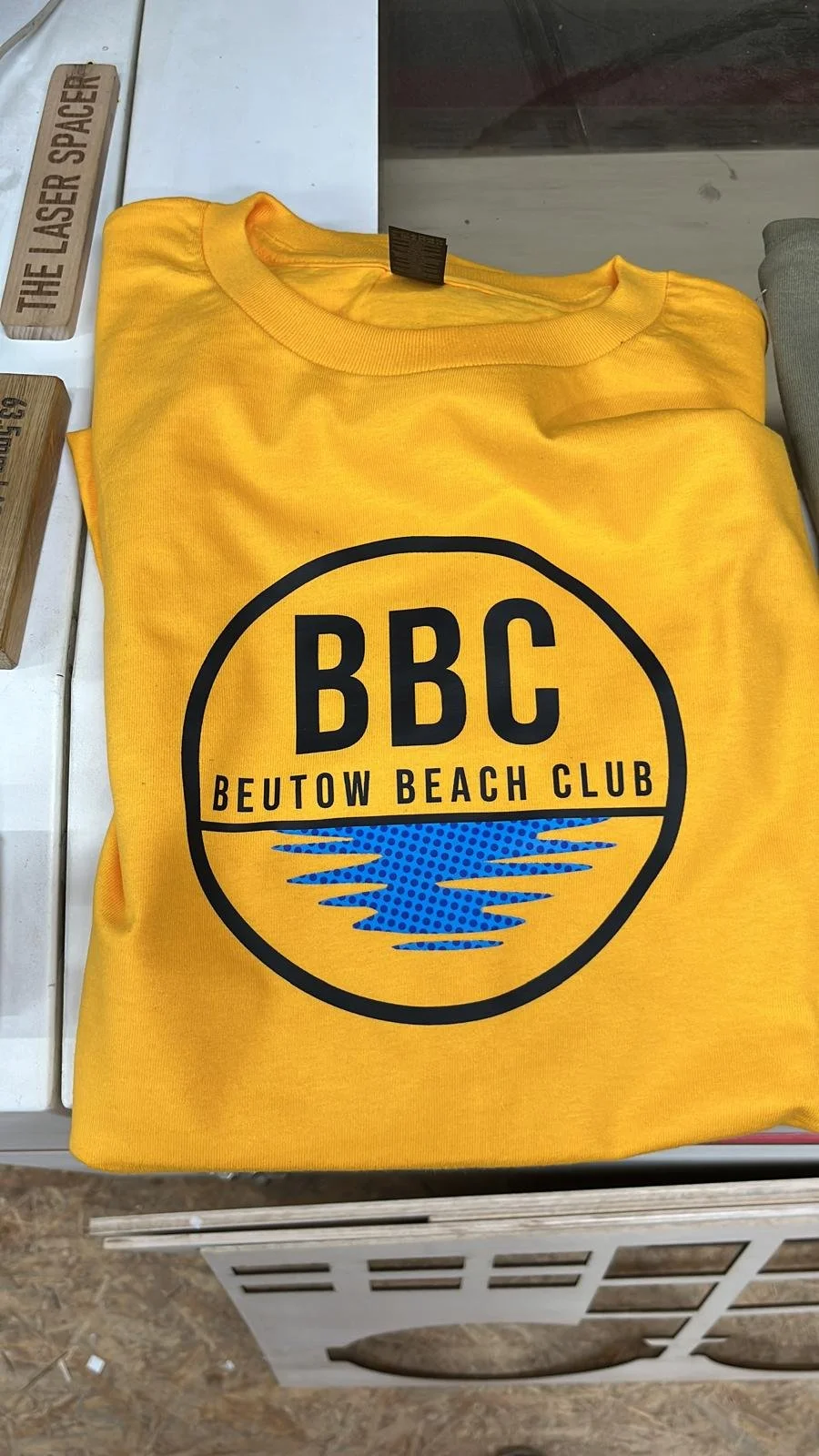 Gelbes T-Shirt mit dem Logo des Beutow Beach Club, das einen blauen Wasserbildschirm und die Buchstaben BBC zeigt.