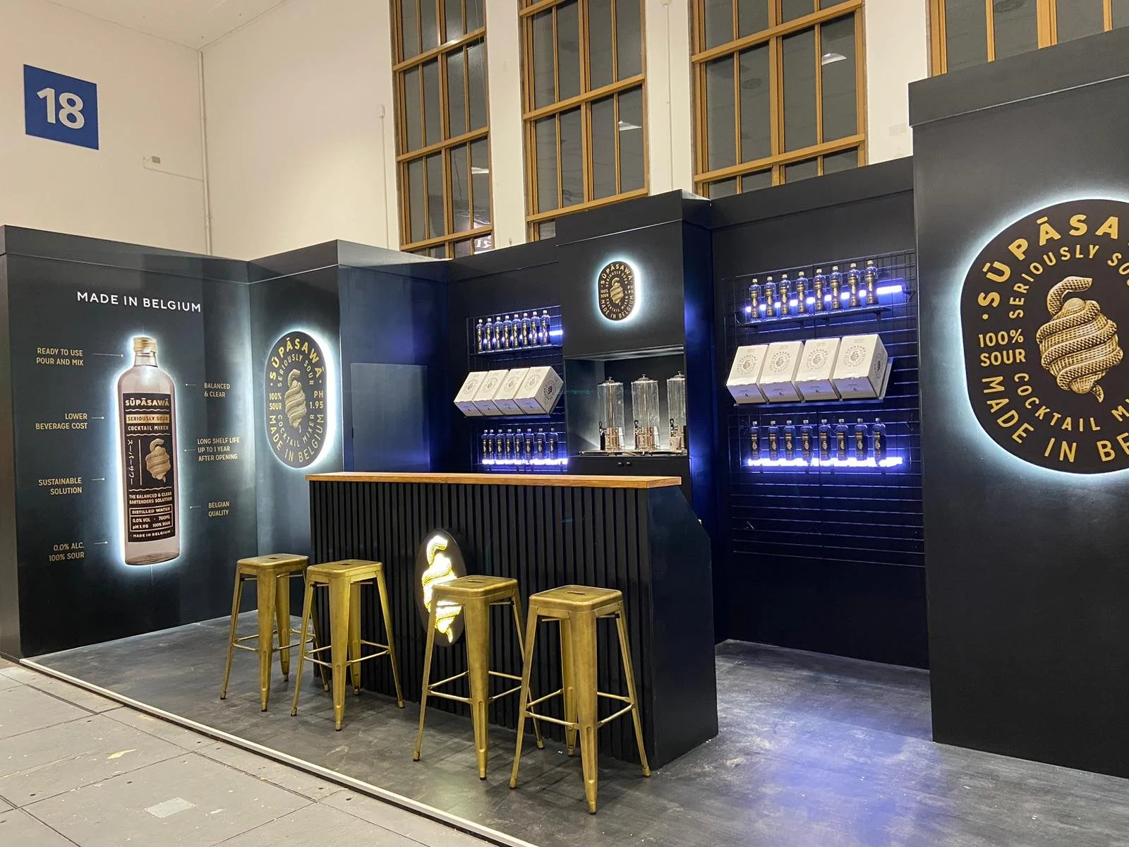 Eine Bar mit schwarzer Wand, beleuchteten Regalen mit Flaschen, weißen Paketen und beigen Barhockern. Das Design ist modern und elegant, mit Beleuchtung in Blau und Weiß.