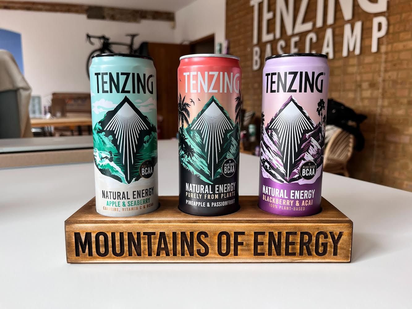 Drei Energydosen mit Tenzing-Logo auf einem Holzständer mit der Aufschrift 'Mountains of Energy' im Hintergrund. Die Dosen haben verschiedene Geschmacksrichtungen: Apfel & Seeb�rrey, Ananas & Passionsfrucht sowie Brombeere & Acai.
