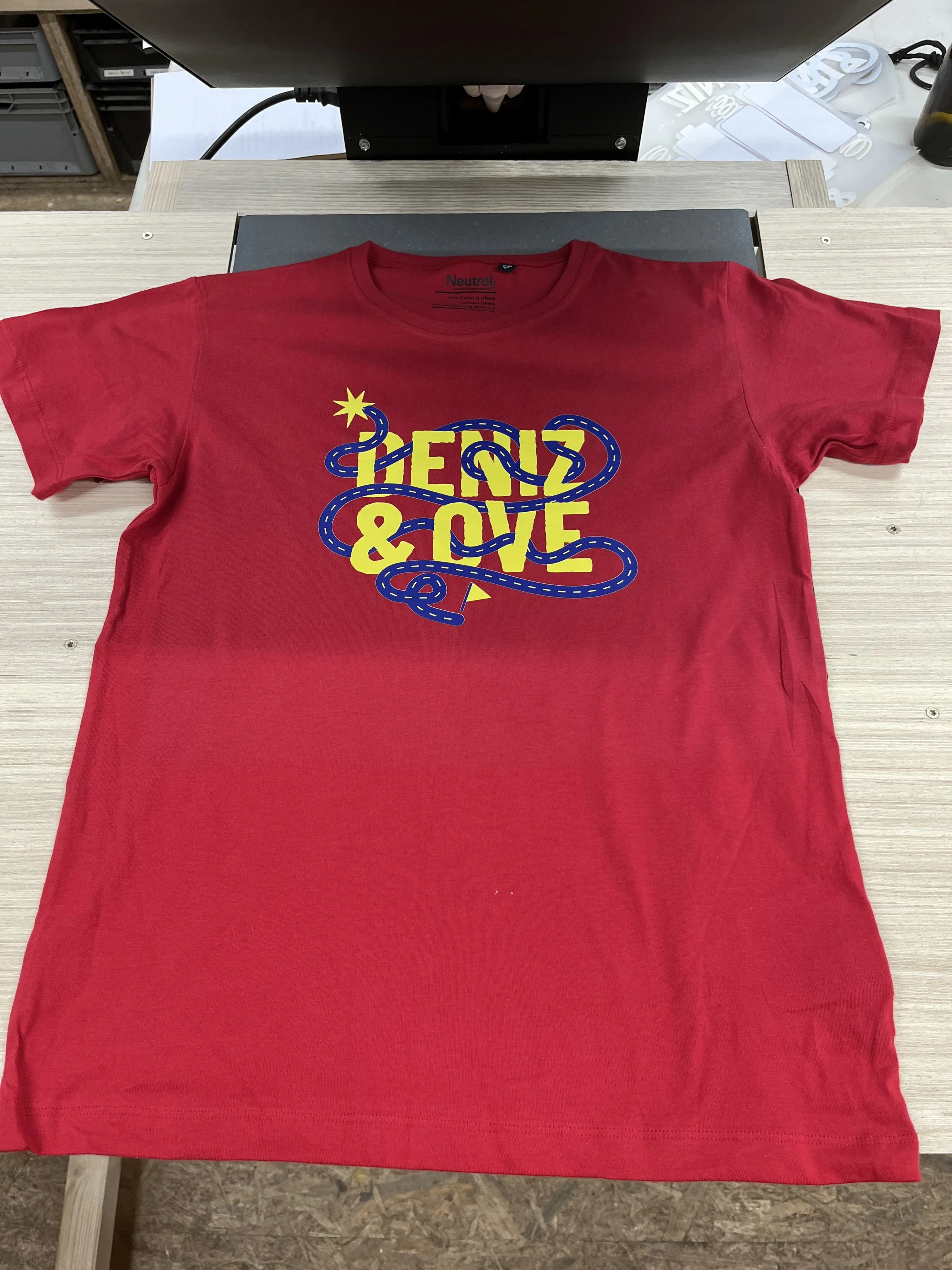 Rotes T-Shirt mit gelb-blauer Aufschrift 'The 2 Love' auf einem Tisch.