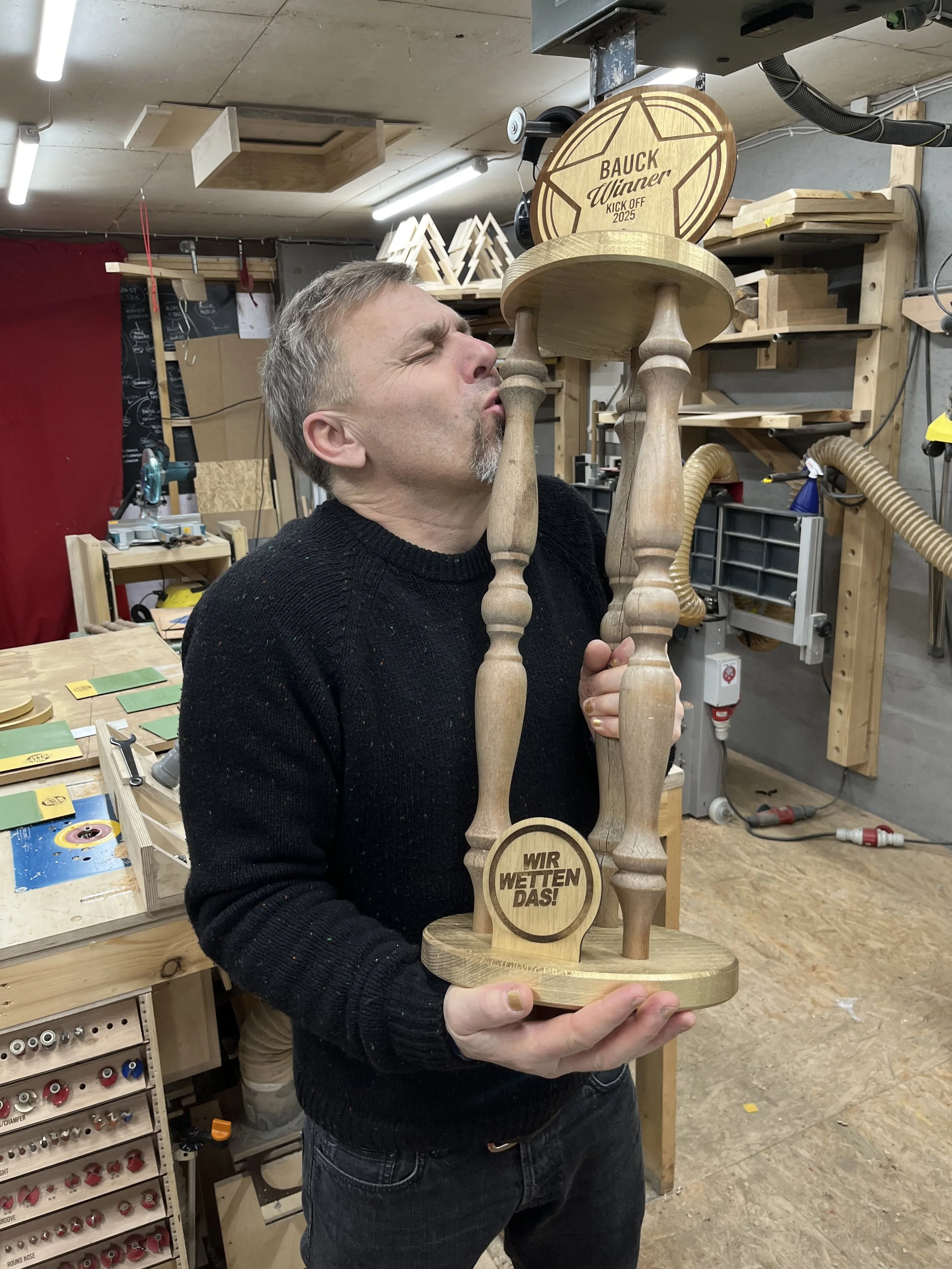 Ein Mann hält einen Holzpokal, auf dem steht 'Wir wetten das!' und eine andere Holzplatte mit 'Bauck Winner Kick Off 2025'. Er steht in einer Werkstatt mit Holzarbeiten und Werkzeugen im Hintergrund.