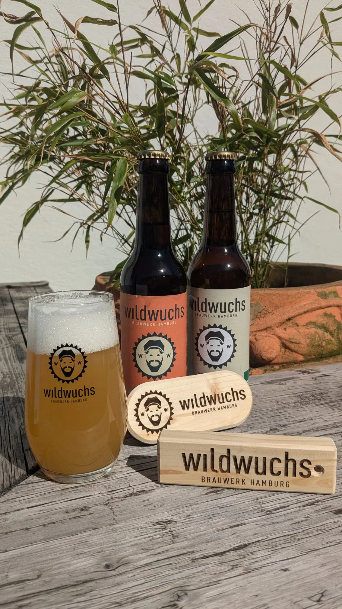 Zwei braune Bierflaschen und ein Bierglas mit hellem Bier auf einem Holztisch. Im Hintergrund eine grüne Pflanze. Die Flaschen und das Glas tragen das Logo und den Namen 'wildwuchs Brauwerk Hamburg'. Es gibt außerdem Holzschilder mit dem Logo und dem
