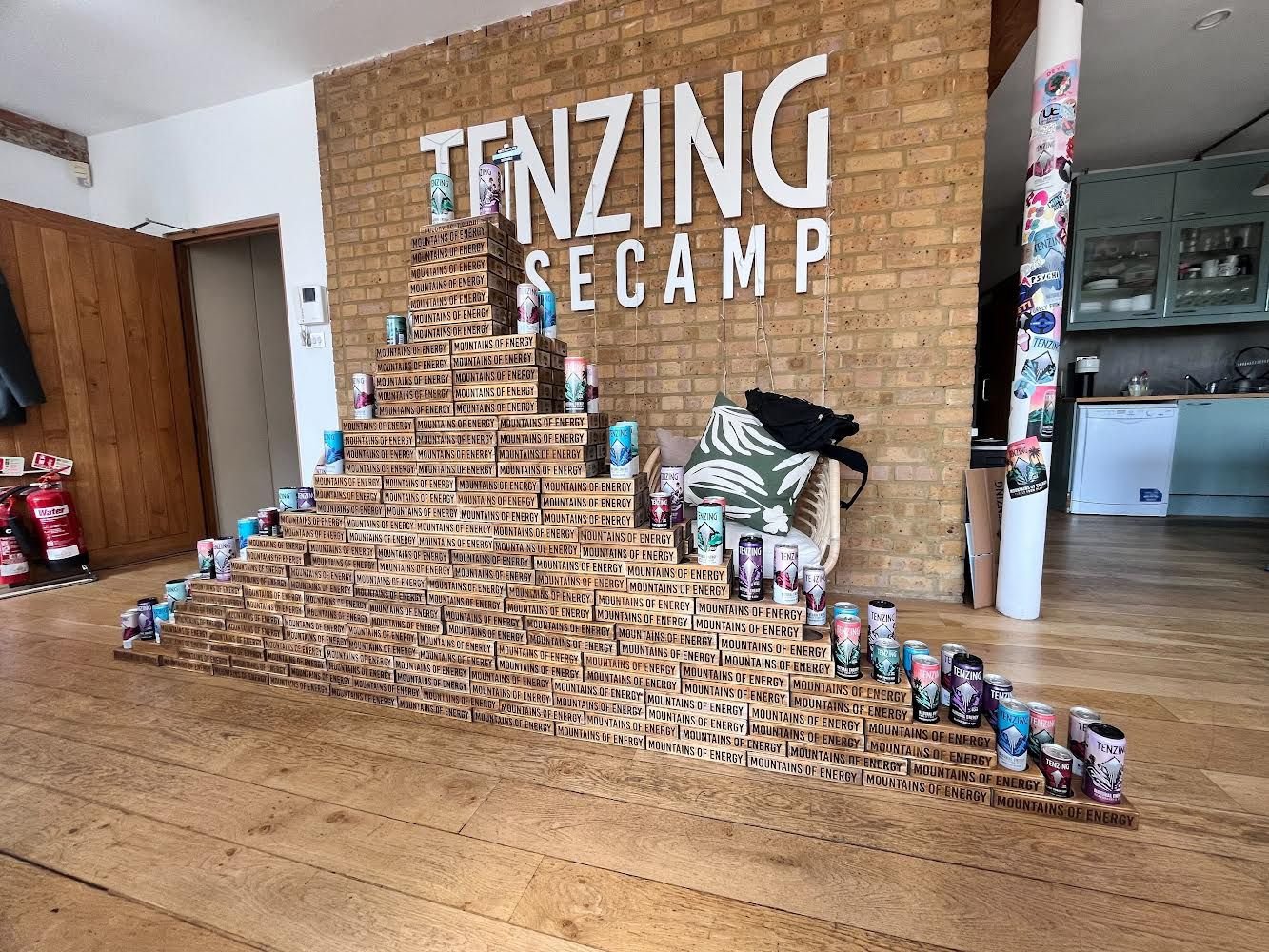 Ein aufgestapeltes Pyramiden aus Energy-Getränkedosen vor einer Ziegelwand mit der Aufschrift "Tenzing Secamp"