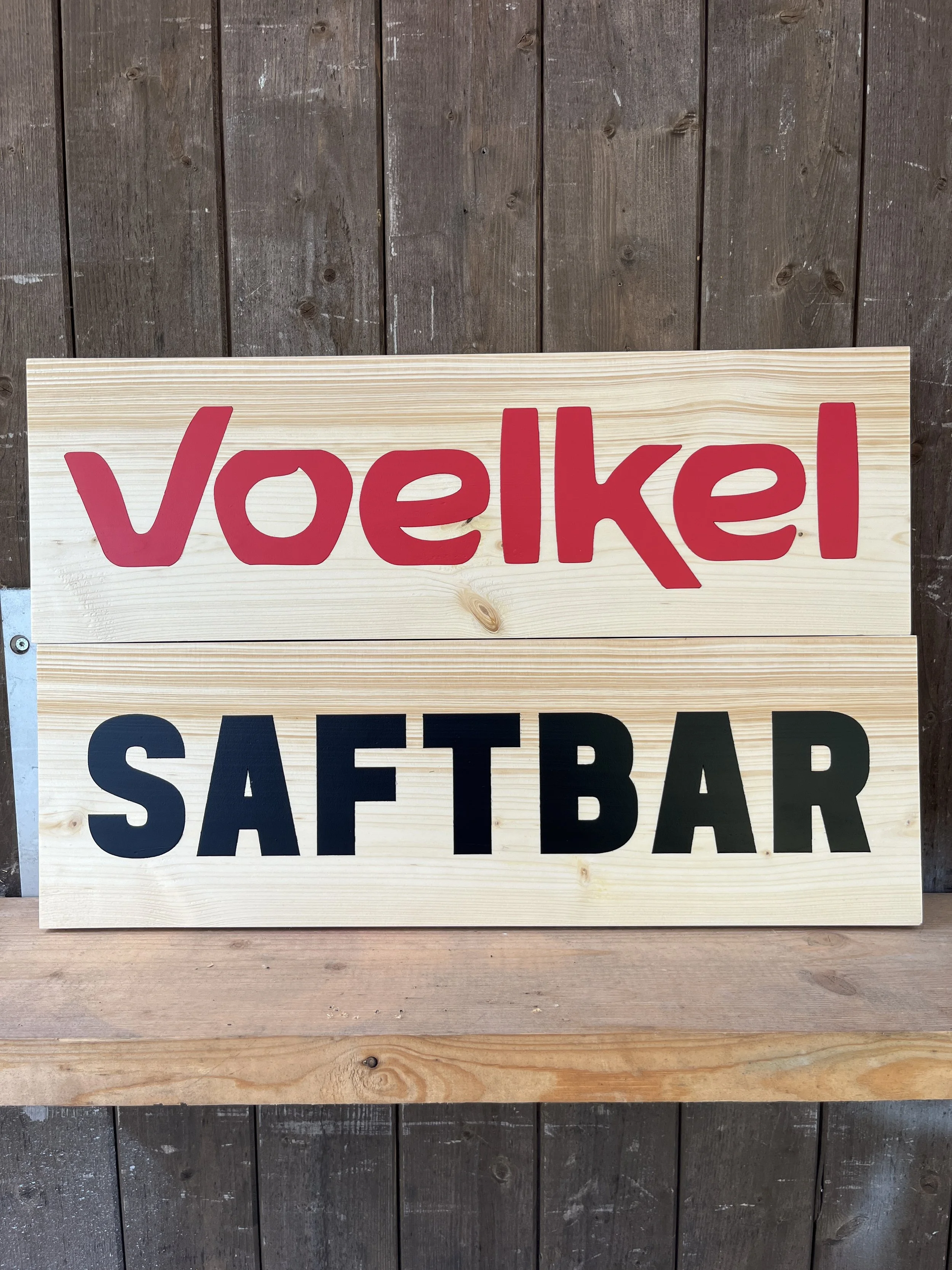 Holzschild mit roter Aufschrift 'Voelkel', darunter ein weiteres Schild mit schwarzer Aufschrift 'SAF BAR', beide auf einem Holzsockel montiert, vor einer holzverkleideten Wand.