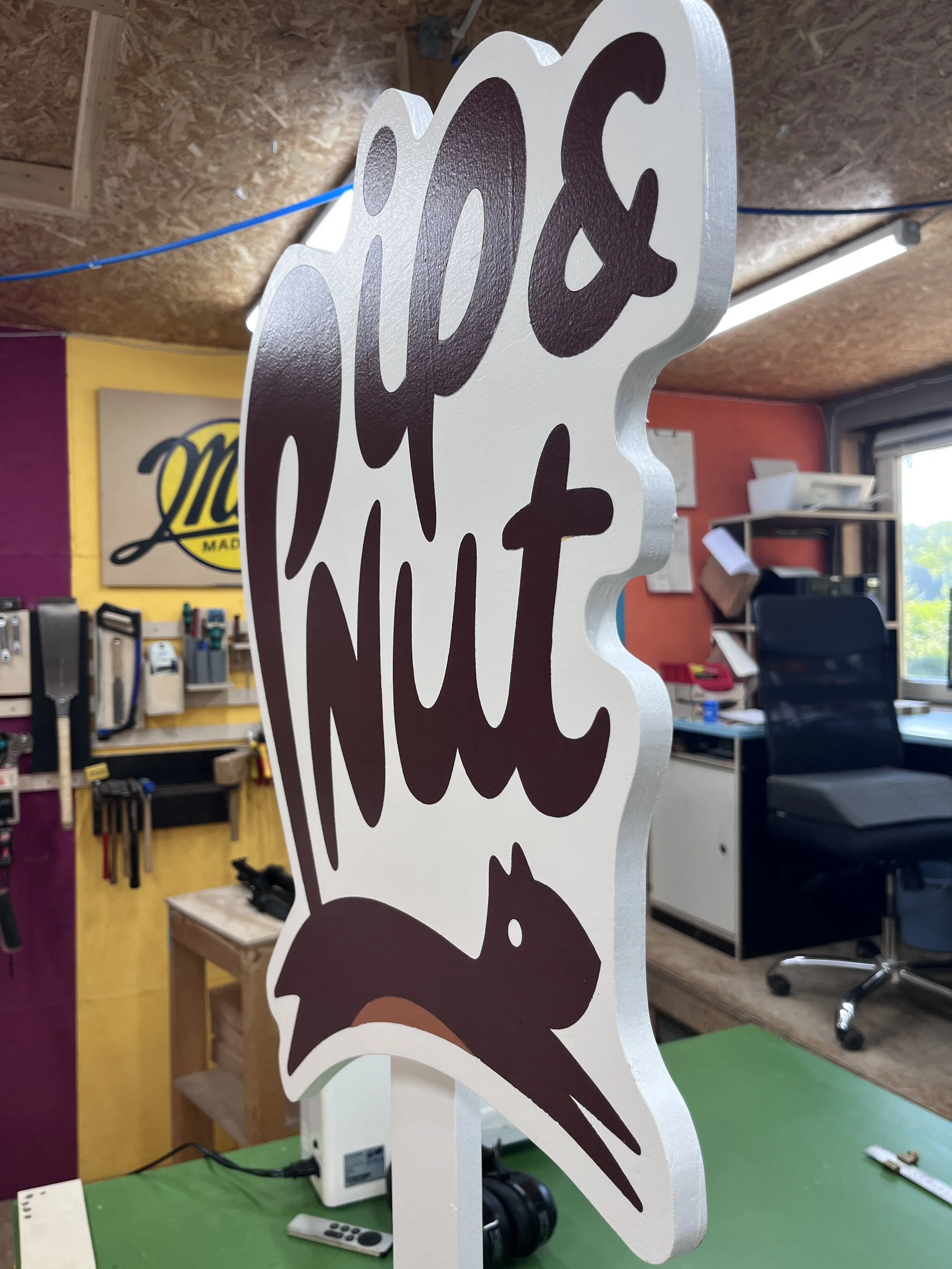 Ein Holzschild in Form eines Hasen mit dem Schriftzug ‚Frohe Ostern‘ darin, auf einem Arbeitstisch in einem Werkraum, im Hintergrund Werkzeuge und Möbel.