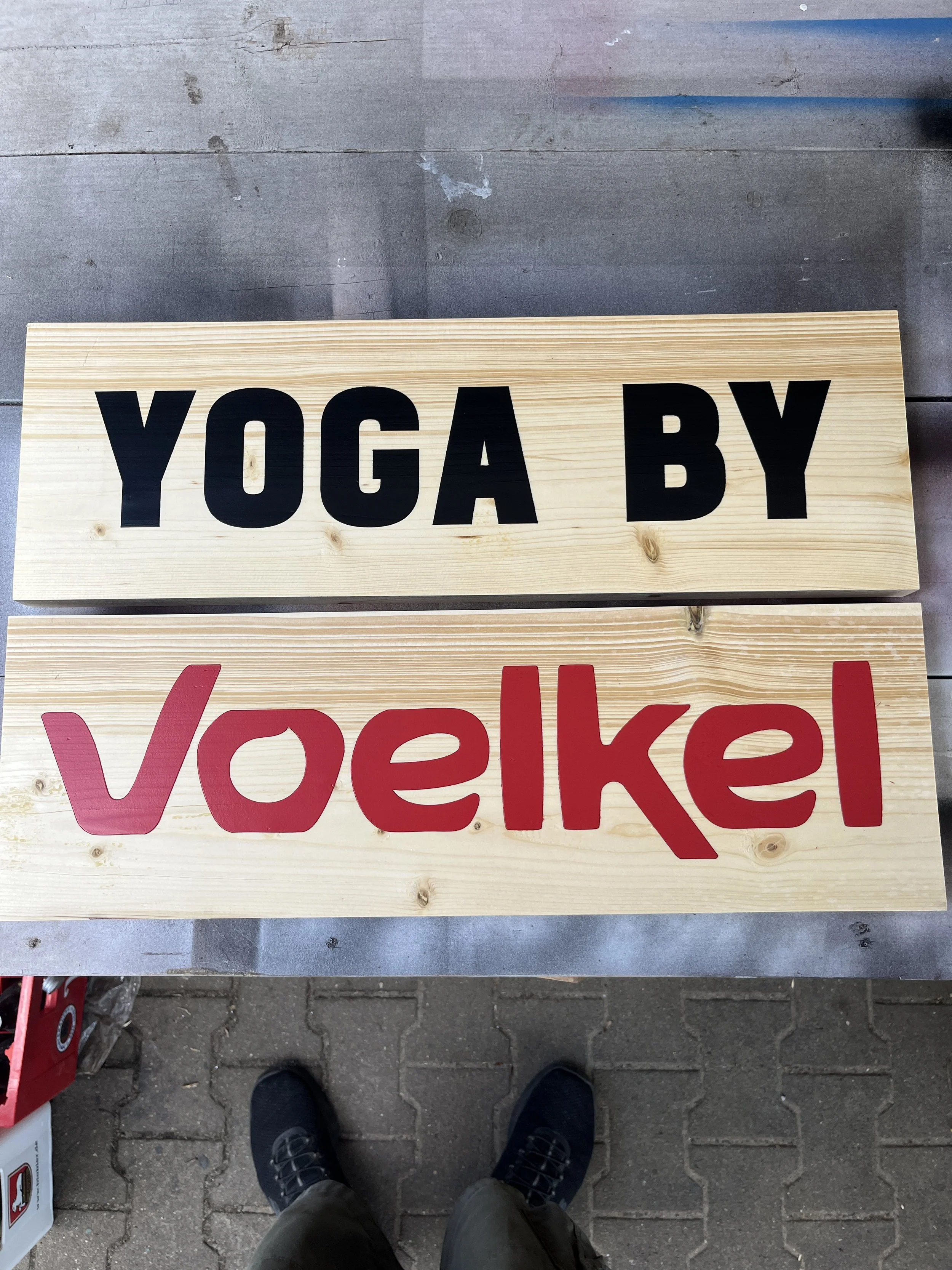 Ein Holzschild mit schwarzen Buchstaben sagt 'YOGA BY' und ein weiteres Schild darunter mit roten Buchstaben sagt 'VoeKe'.