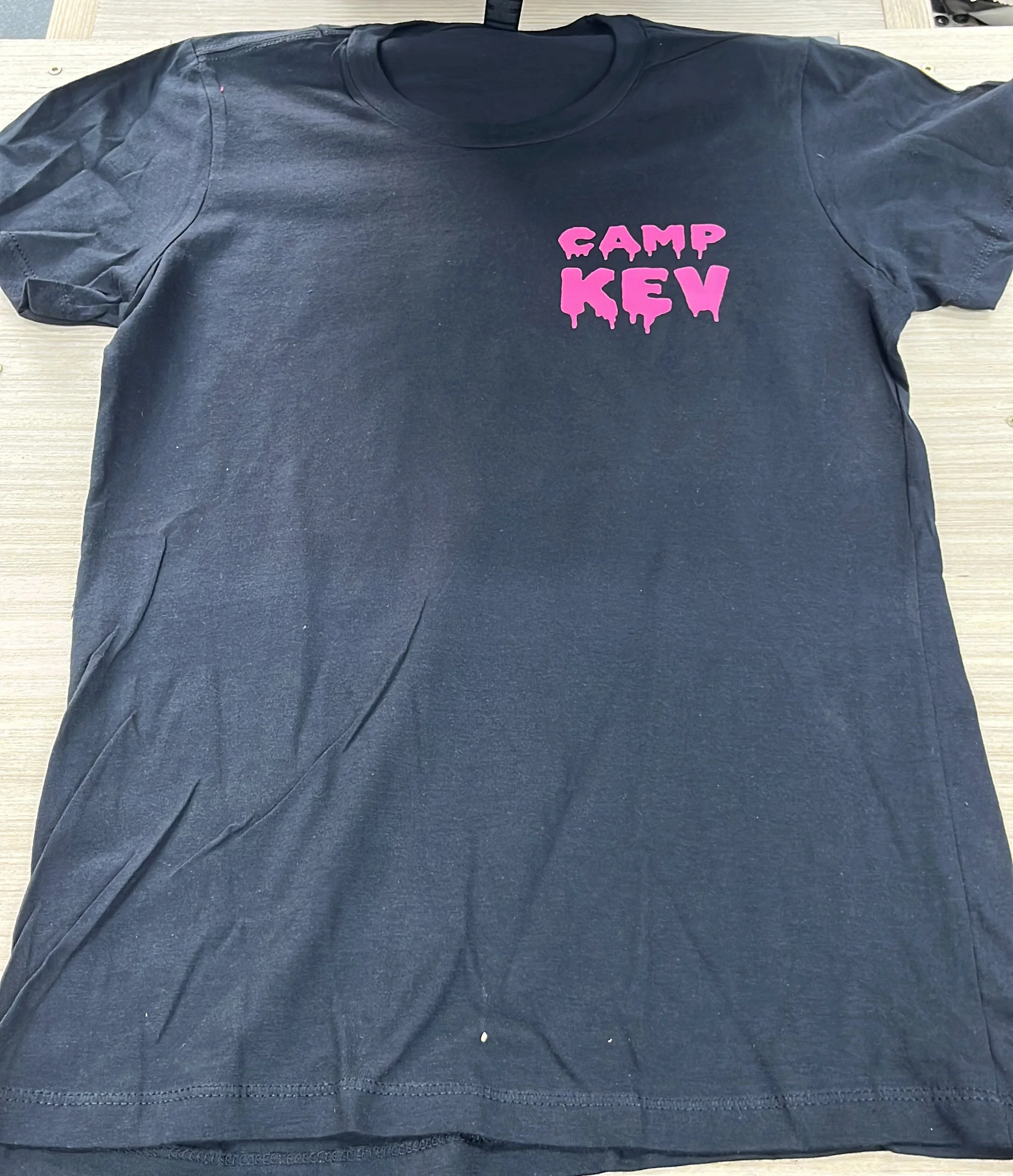 Schwarzes T-Shirt mit pinker Aufschrift "CAMP KEW" auf einer Holzplatte liegend.