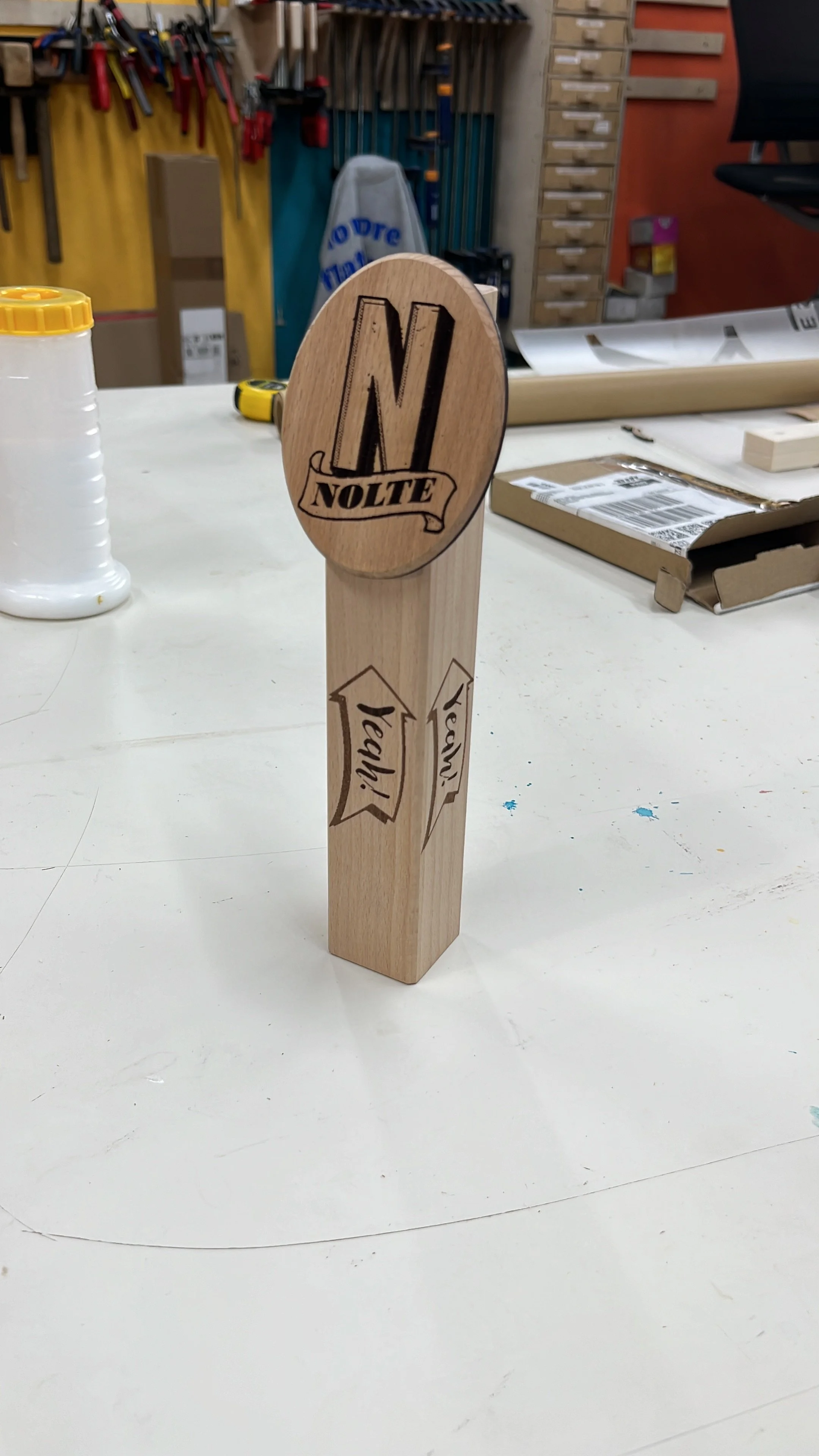 Holzwerkzeug mit graviertem Logo "NL NOFTE" und Sprüchen "Yea!" und "Yes!" in einer Werkstatt. Hintergrund mit Werkzeugen und Werkbänken.