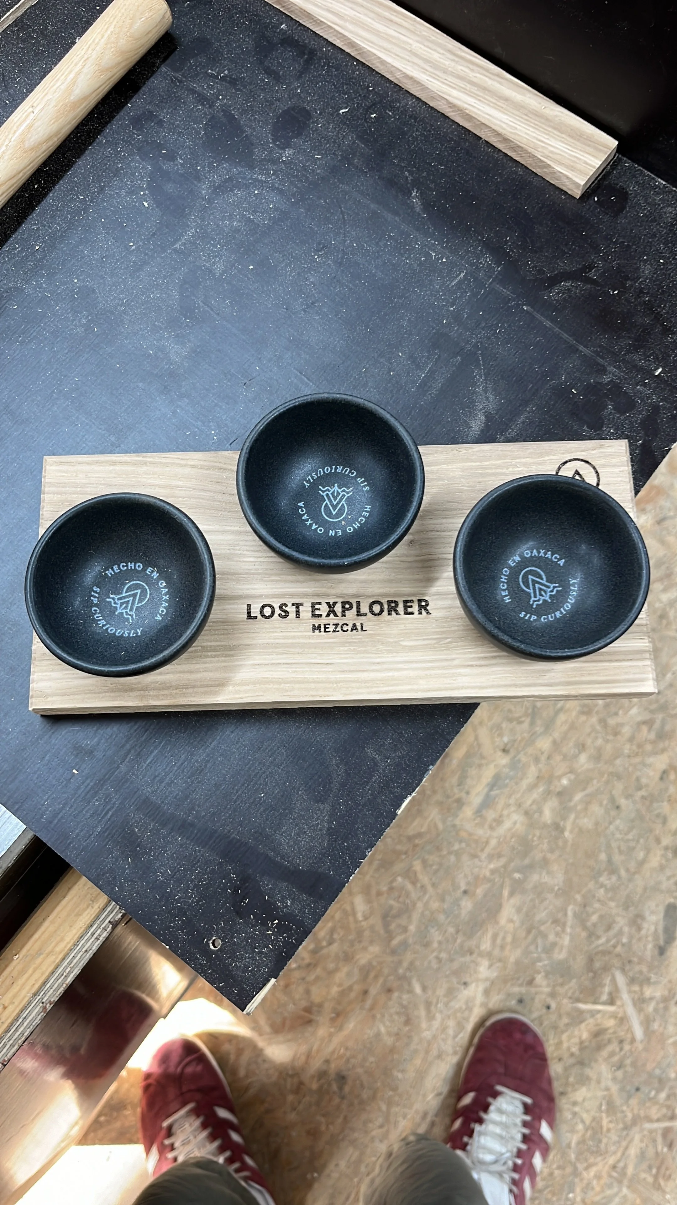 Drei schwarze Keramikschalen stehen auf einem Holzbrett mit der Aufschrift 'Lost Explorer Mezcal'. Die Schalen tragen den Text 'Hecho en Oaxaca, Sip Curiously' und haben ein Logo. Der Boden ist aus Holzplatten, und es sind Füße von Personen mit Sneak