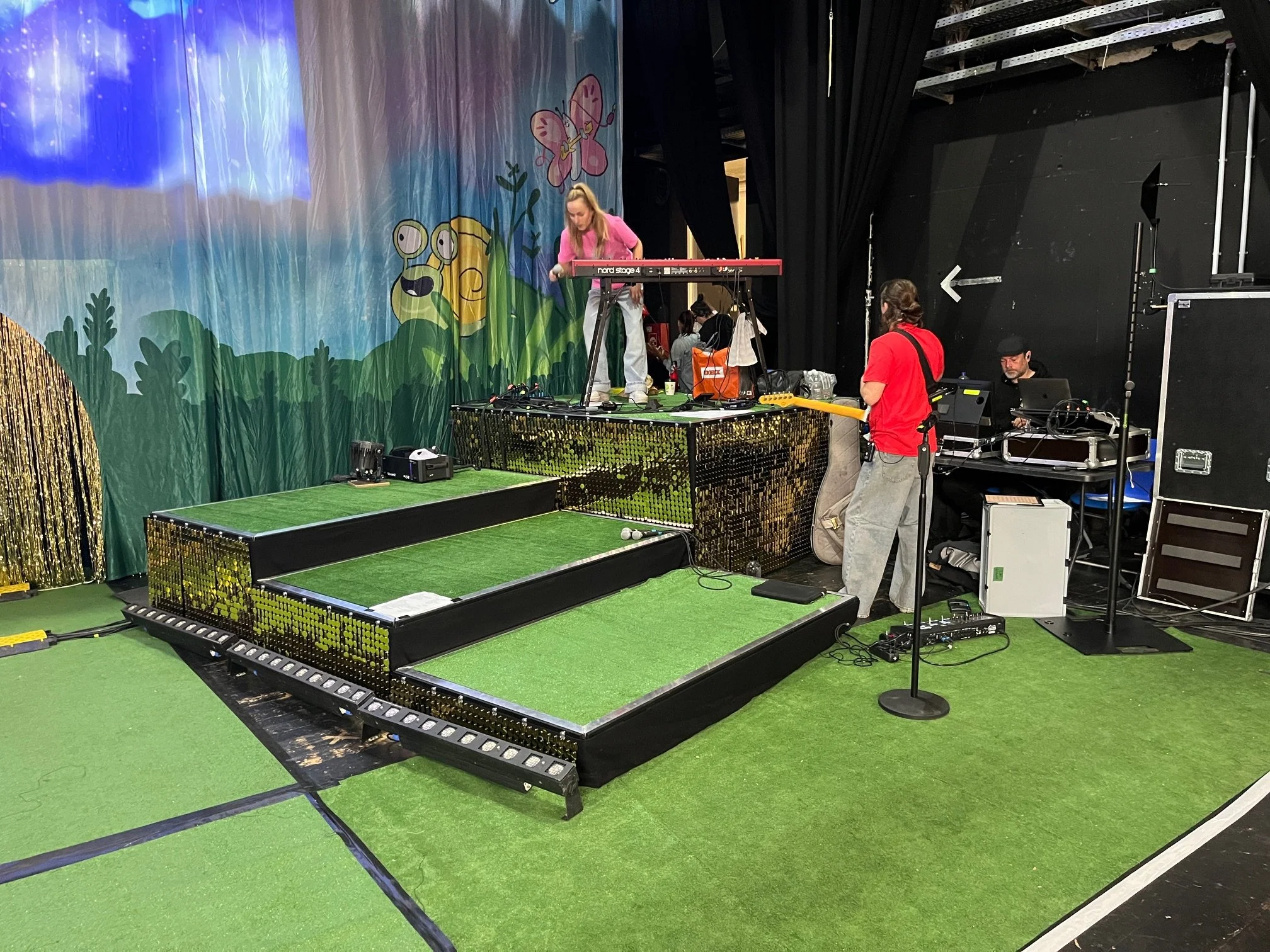 Bühne mit gemalten Tieren im Theater, weiße Menschen, technische Ausrüstung und grüne Teppich am Boden, ein Techniker trägt Rot, einem Musiker in Rosa und Grau bei der Vorbereitung für eine Aufführung.