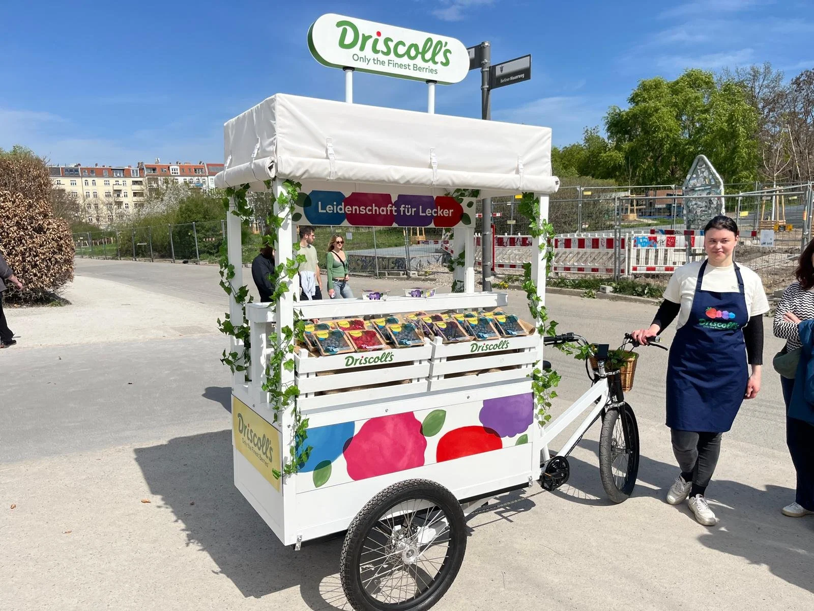 Mobiler Verkaufsstand für Beeren mit dem Namen Driscoll's, geschmückt mit grünen Blättern, mit einer Verkäuferin im blauen Apron, die neben einem Fahrrad steht.