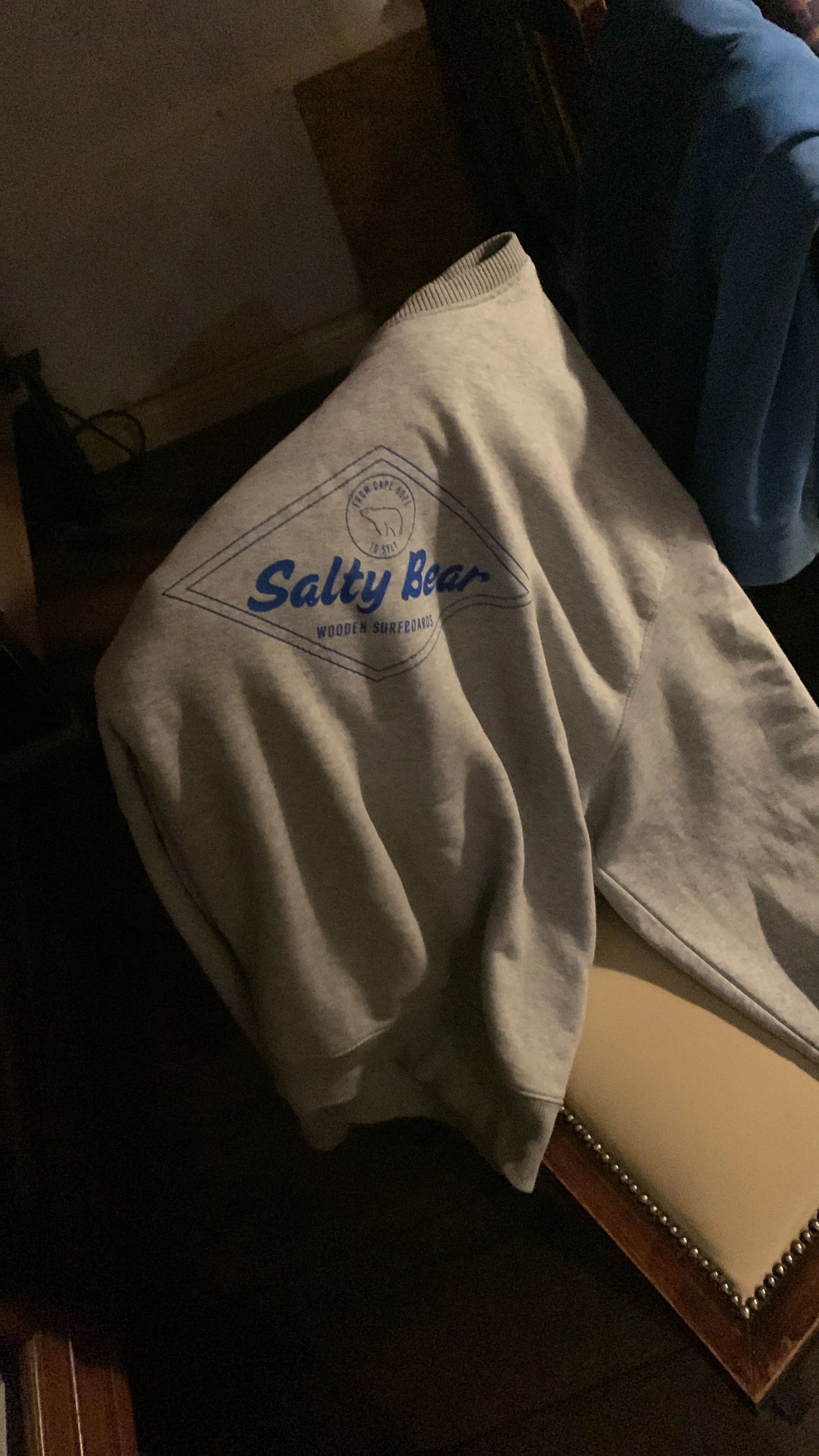 Ein graues Sweatshirt mit blauer Aufschrift "Salty Bear" und einem Bärenlogo liegt auf einem Stuhl.