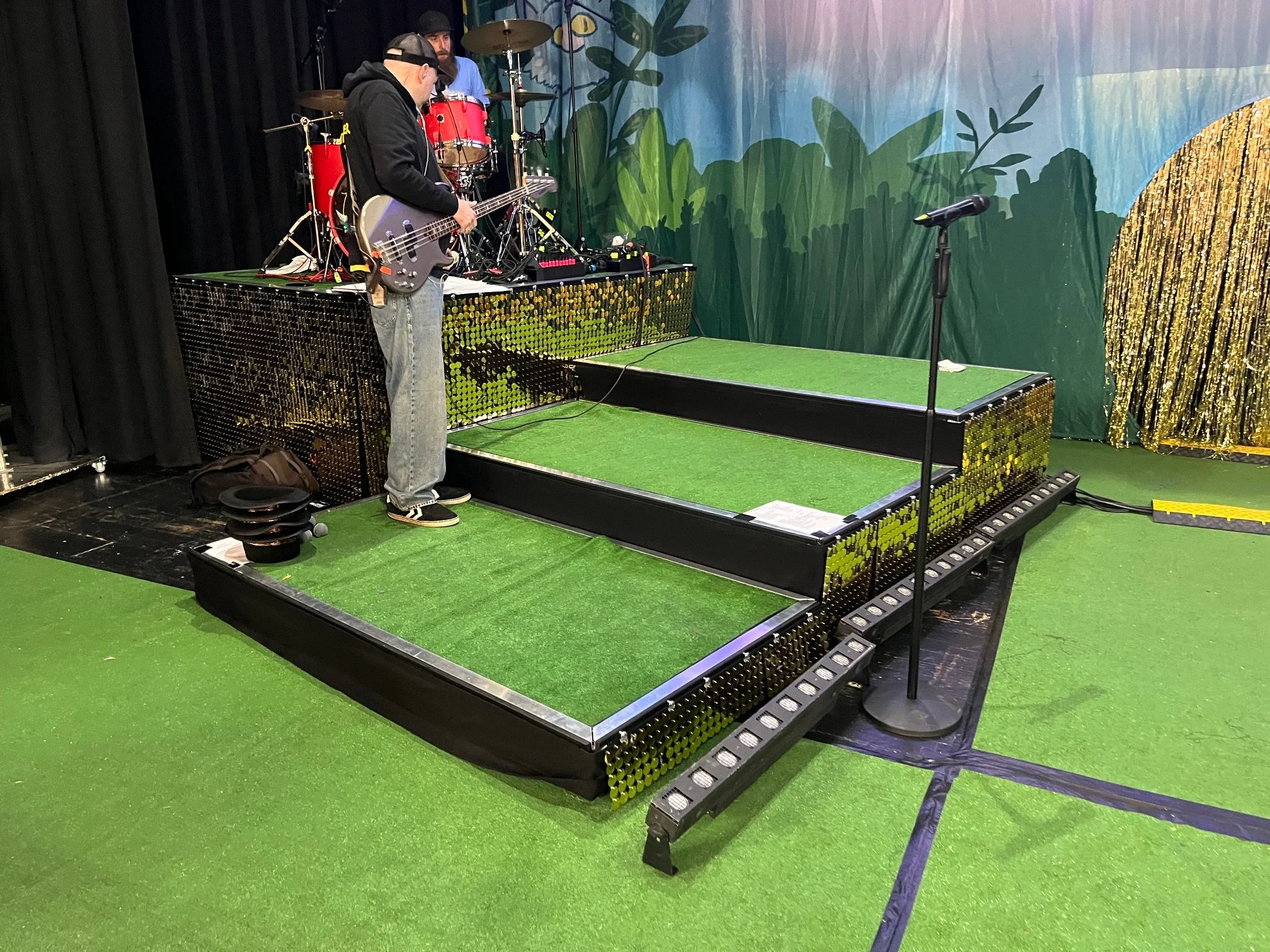 Bühne für eine Musikperformance mit einem Bassisten und einem Schlagzeuger, grüne Teppiche und Dekorationen im Hintergrund.