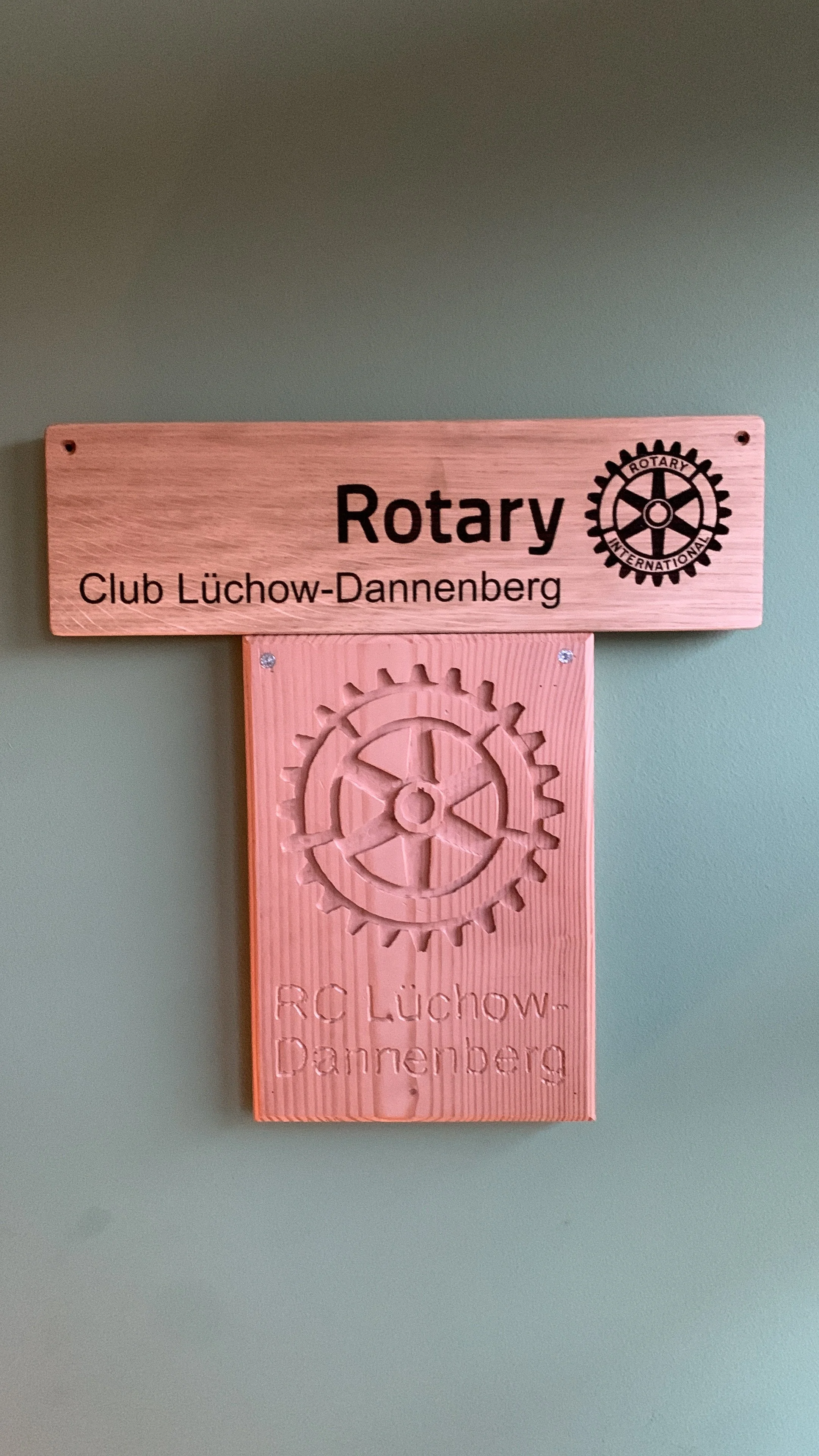 Ein Holzschild mit dem Rotary-Logo und dem Text 'Club Lüchow-Dannenberg' hängt an einer grünen Wand.