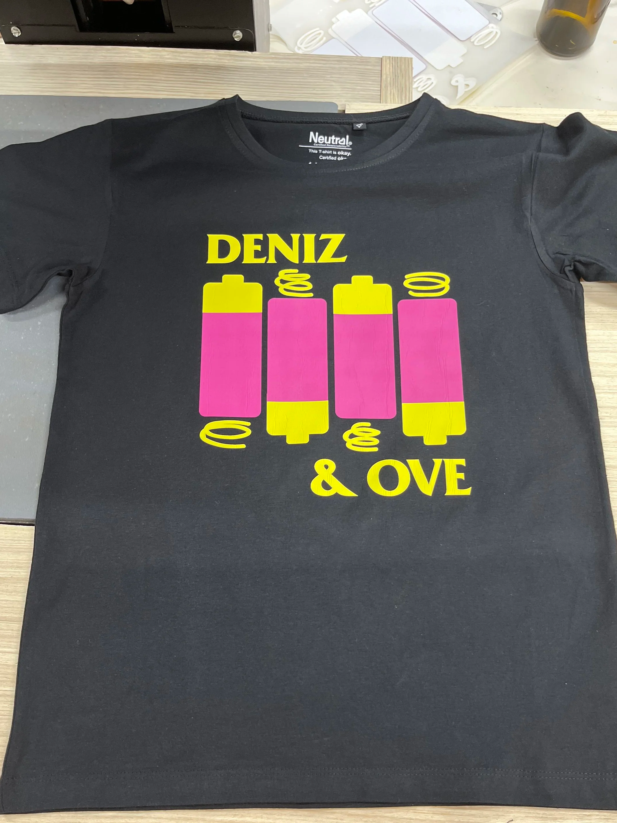 Schwarzes T-Shirt mit buntem Druck, der vier Batterien und den Worten 'DENİZ & OVE' zeigt.