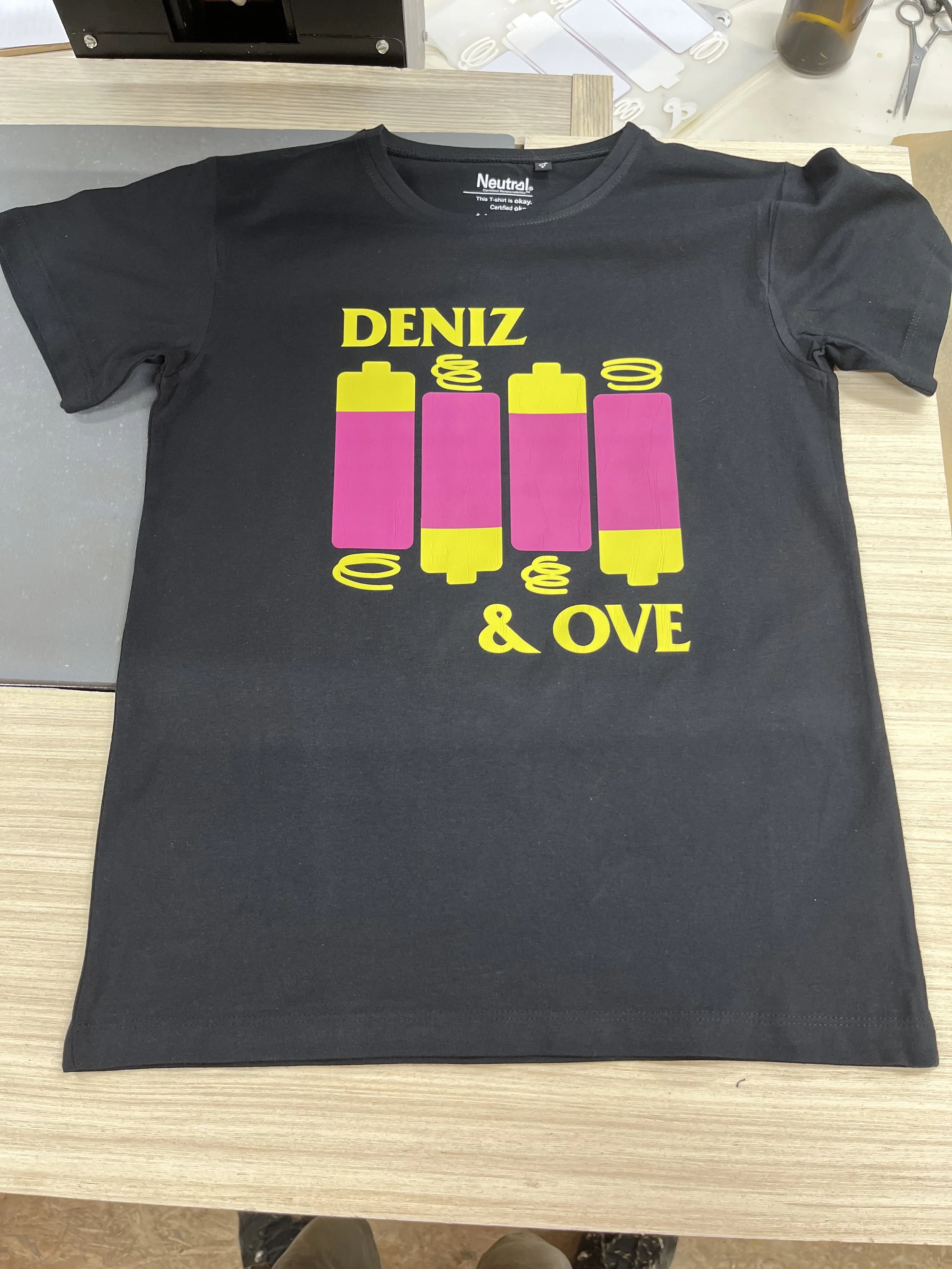 Schwarzes T-Shirt mit gelben und rosa grafischen Elementen und gelbem Text, darauf steht 'DENIZ & OVE'.