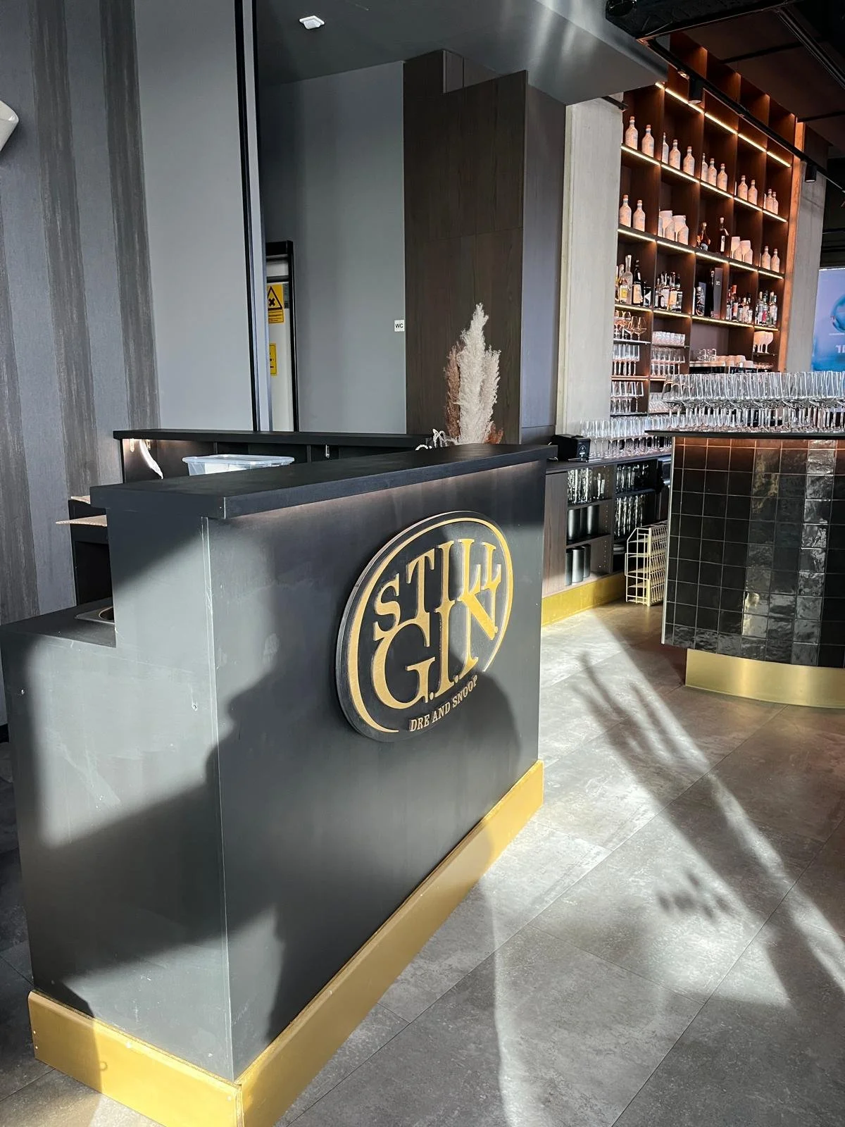 Bar mit einer schwarzen Theke, auf der ein runder Schriftzug mit dem Wort "STIL GIN" in Gold steht, darunter der Satz "DREAM AND SHOOT". Im Hintergrund sind Regale mit Flaschen und Gläsern, die die Bar-Kulisse vervollständigen.