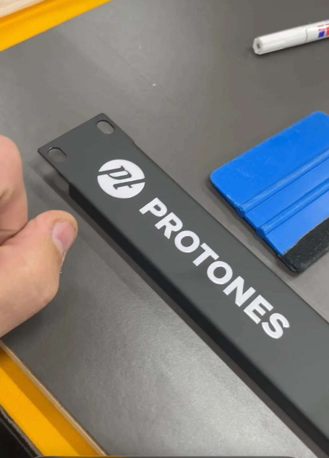 Hand hält ein schwarzes Schild mit dem HP-Logo und der Aufschrift 'PROTONES' auf einem Tisch, neben einem blauen Gegenstand und einem weißen Marker.