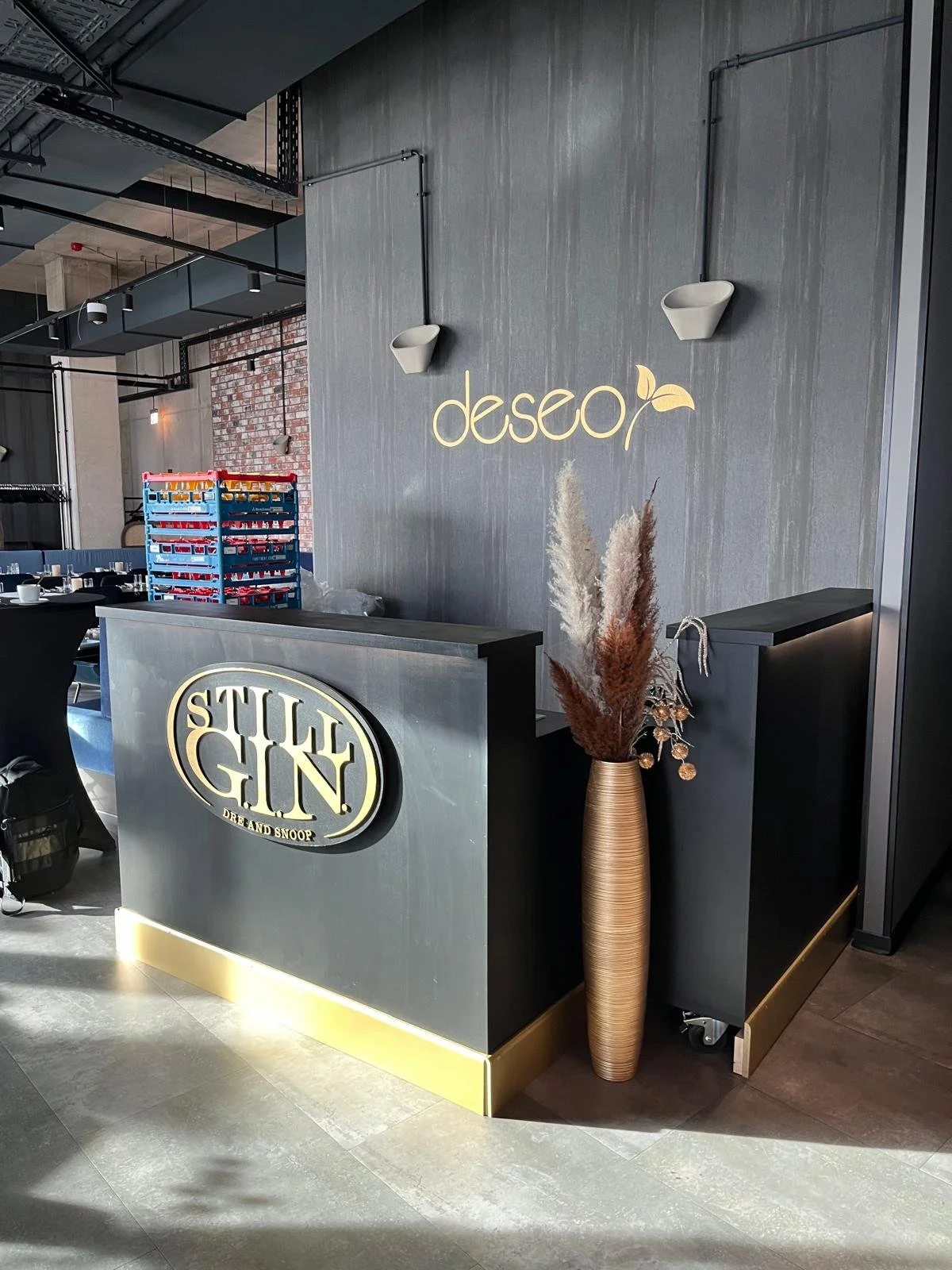 Innenaufnahme eines Restaurants oder Cafés mit der Beschriftung 'deseo', moderner Dekoration und einer Theke mit einem Logo, vertikaler Vase mit Trockenblumen.