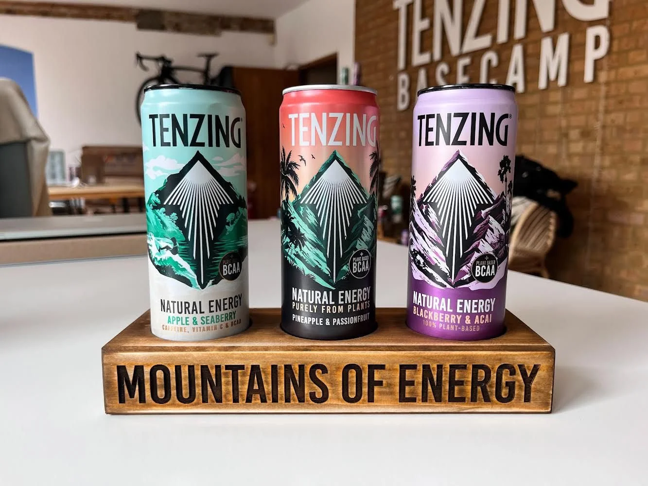 Drei Tenzing Energy-Getränkedosen auf einem Holzständer mit der Aufschrift "Mountains of Energy". Die Dosen haben unterschiedliche Designs und Geschmacksrichtungen: Apfel & Seaberry, Ananas & Passionsfrucht, Brombeere & Acai.