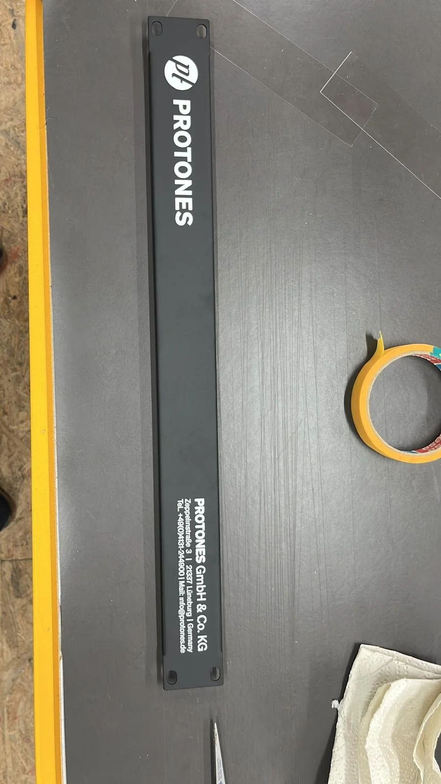 Schwarzes Metallstück mit weißen Aufschriften und einem Logo, vermutlich ein Name und Kontaktdaten, auf einem grauen Tisch.