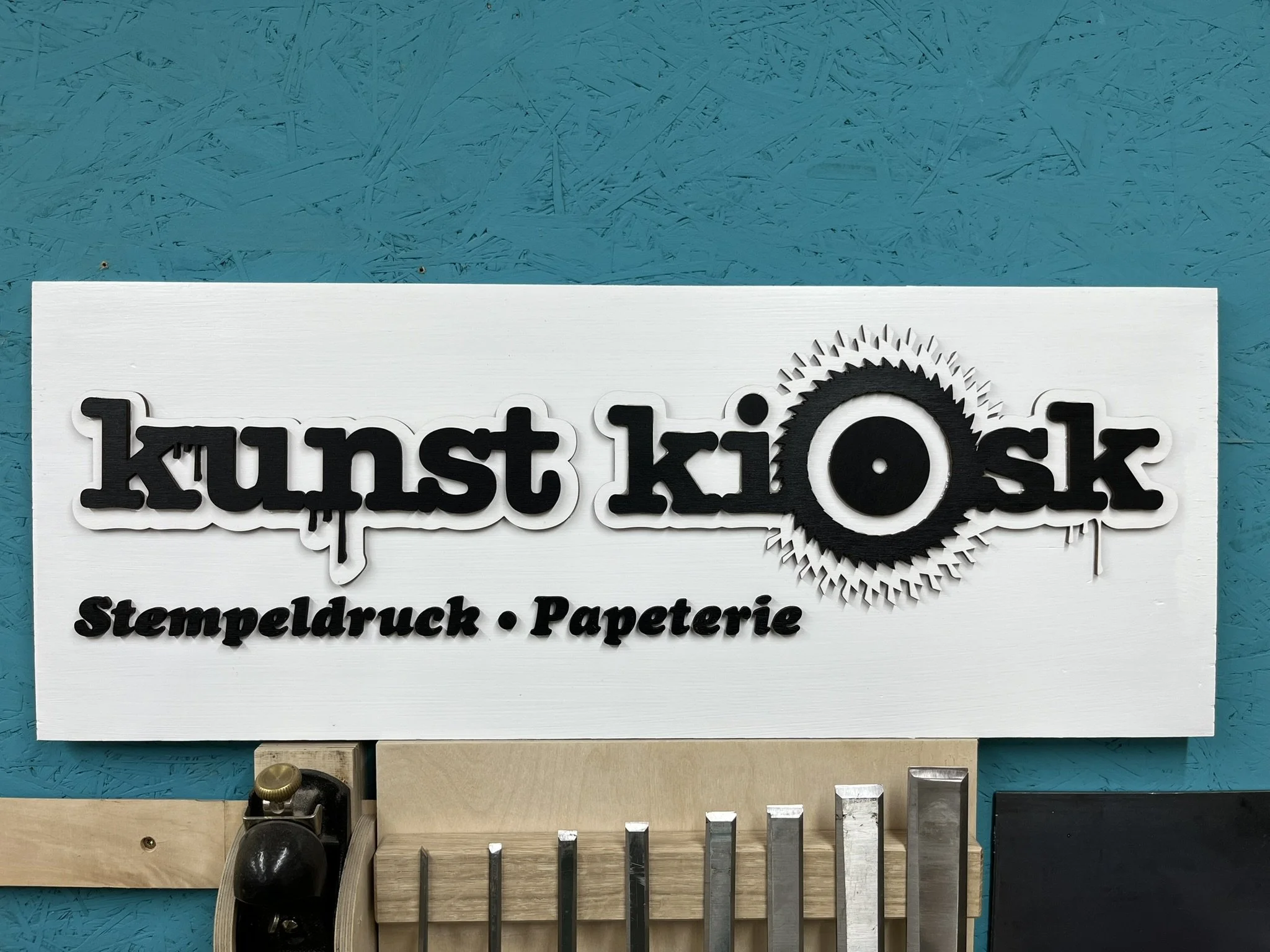 Buchstaben in schwarzer und weißer Farbe, die das Wort 'kunst.kiosk' auf einer weißen Platte anzeigen, darunter befinden sich Begriffe wie 'Stempeldruck' und 'Papeterie'. Im Hintergrund eine blaue Wand mit strukturierter Oberfläche.