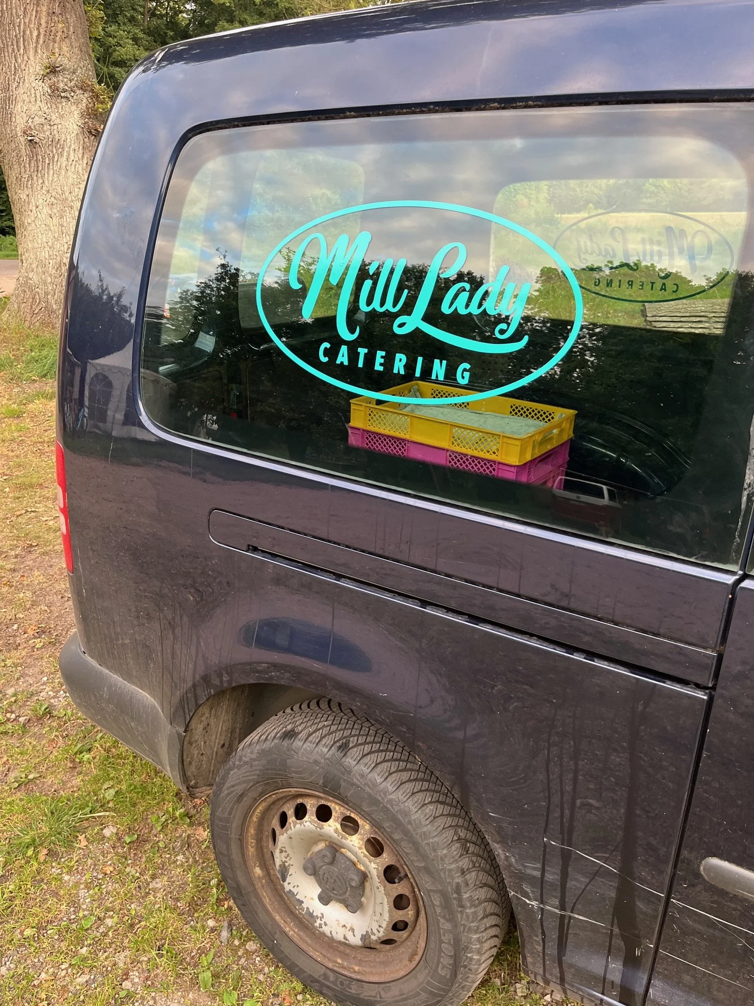 Die Rückseite eines schwarzen Vans mit einem auffälligen Abzeichen für 'MillLady Catering' auf der Heckscheibe, darunter gelbe und pinke Plastikbehälter im Inneren, auf einem grasbewachsenen Feld neben einem Baum.