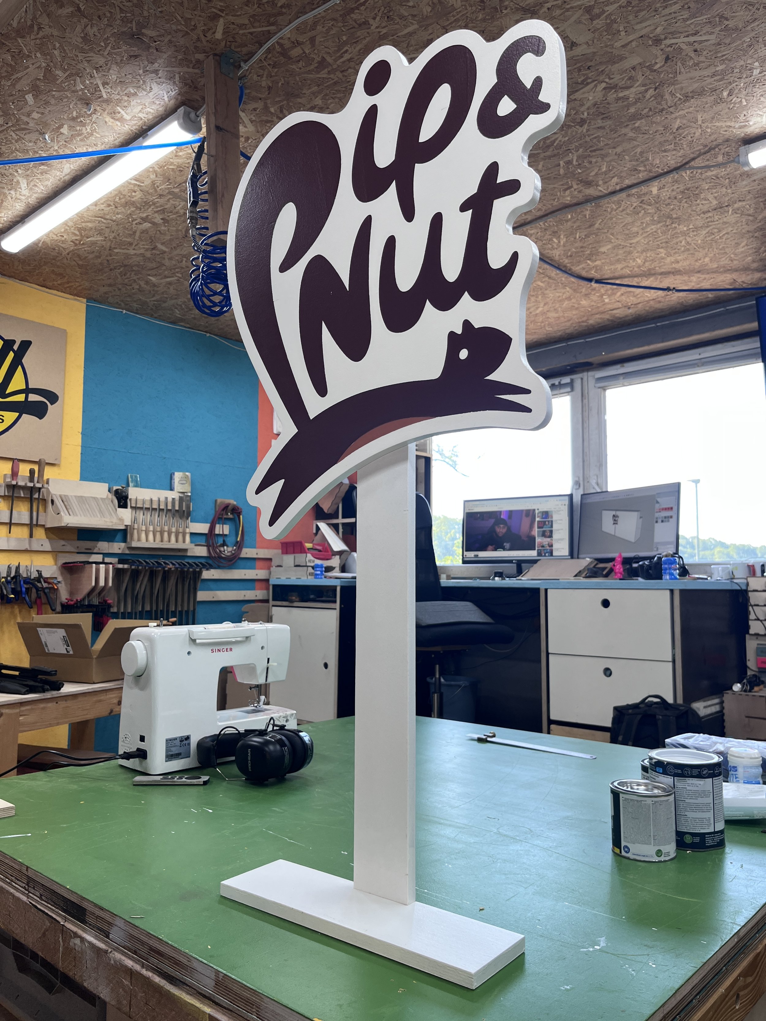 Ein bemalter Holzschild mit der Aufschrift 'Papier mit' und einer stilisierten Ente, steht auf einem grünen Arbeitstisch in einem Werkraum mit Werkzeugen und Computern im Hintergrund.