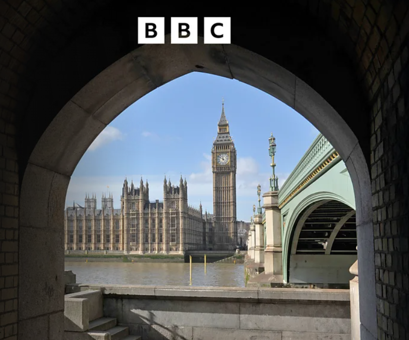 View of Big Ben clock tower and the Palace of Westminster as seen through an archway near the Thames River, with Westminster Bridge on the right.