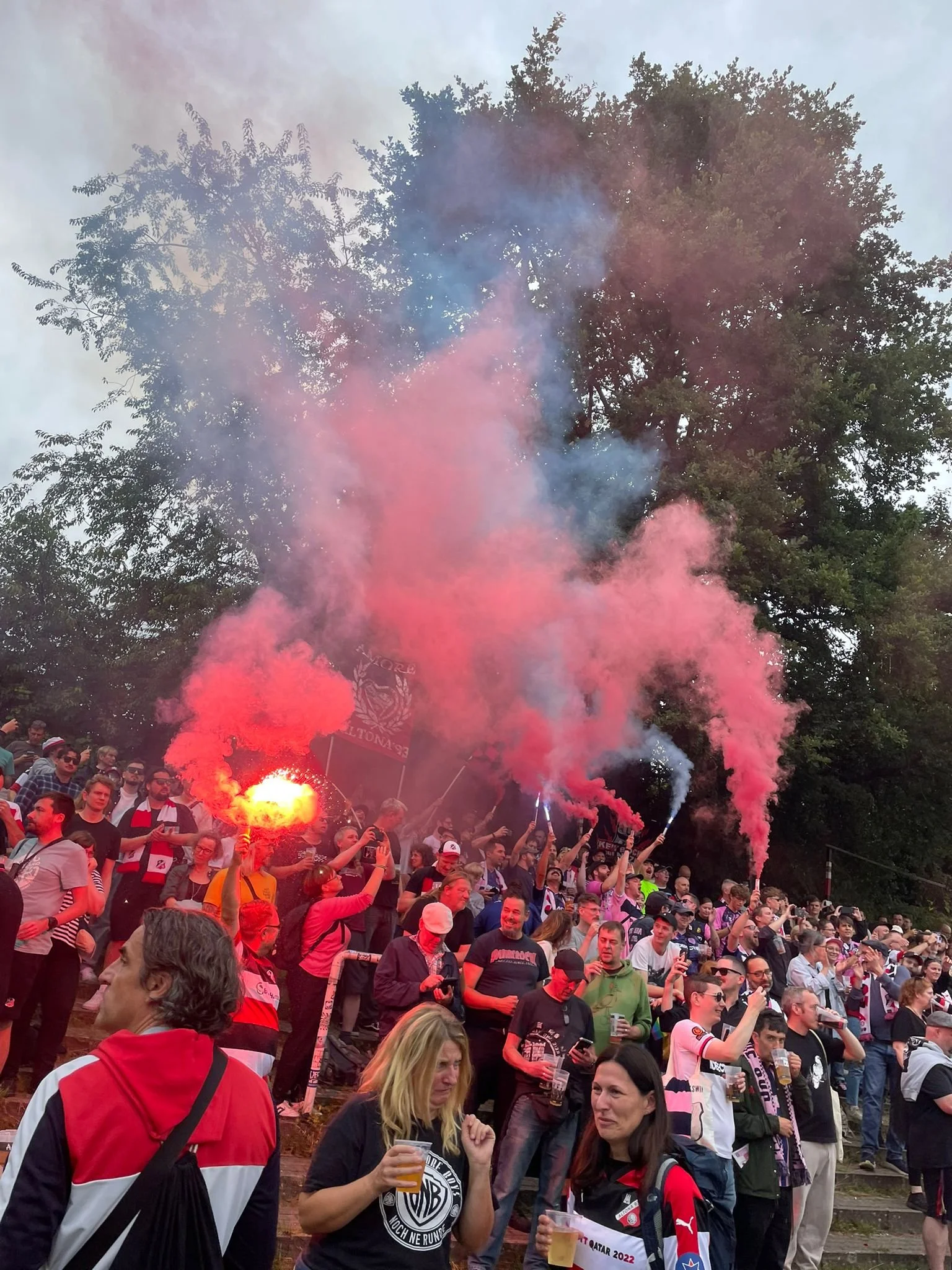 A crowded outdoor event with many people holding up smoke flares emitting pink and blue smoke. Some people are taking photos, holding drinks, and wearing sports jerseys and scarves, suggesting they are fans attending a sporting event or celebration.