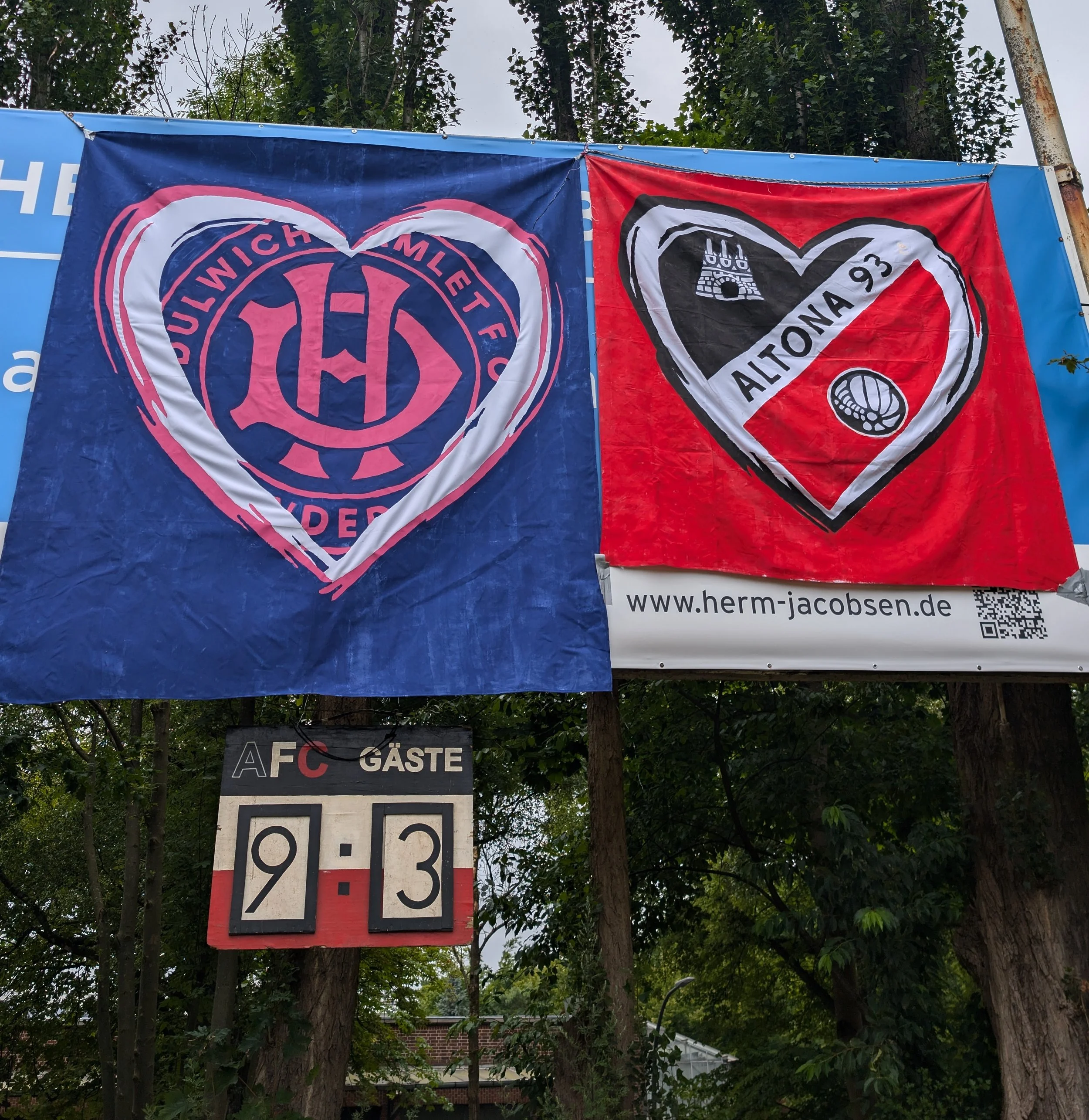 Two flags hanging between trees, one with a blue background and heart-shaped emblem with text, the other red with a black outline and emblem, and a URL. Below, a road sign with the numbers 9-3 and the word GÄSTE.