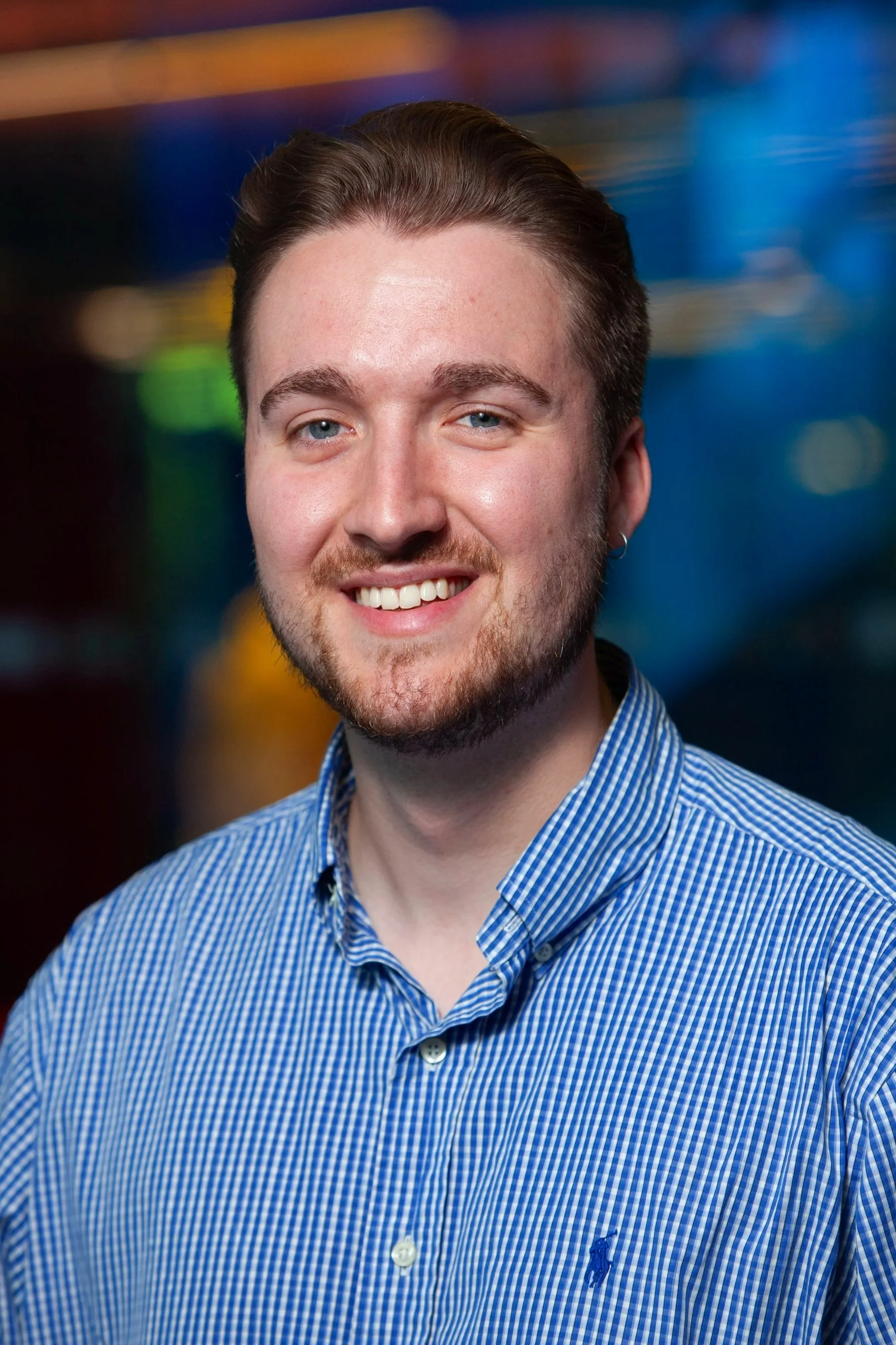 Close-up portrait of a young man with light skin, short brown hair, blue eyes, and a light beard, smiling and wearing a blue checkered button-up shirt with a small logo of a polo player on the chest.